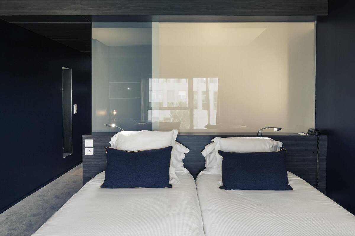 A modern hotel room with a large mirror, two beds with white and dark blue bedding, a desk with a lamp, and a view of the outside through the window.