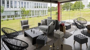 A modern lounge area with a sofa, chairs, and a coffee table, featuring large windows with a view of a building and a grassy area. - Hôtel Mercure Nantes Ouest Saint-Herblain Zenith (Saint-Herblain)