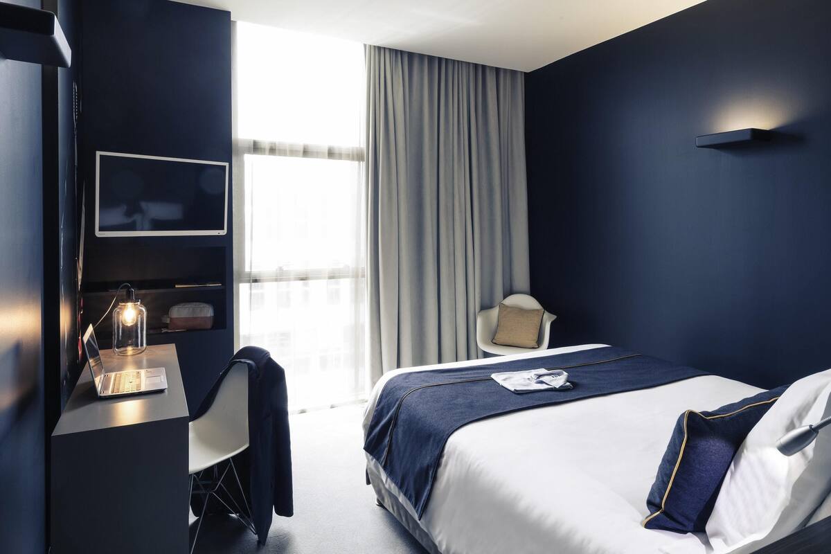 Ein modernes Hotelzimmer mit einem Bett, einem Schreibtisch mit einem Laptop, einem Stuhl, einem Fernseher und einem großen Fenster mit Vorhängen.