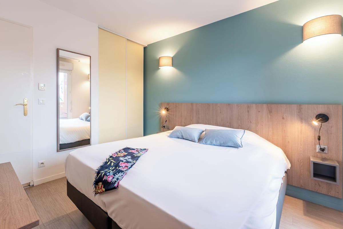 A hotel room with a bed, a mirror, a bedside table, and a wall-mounted lamp.