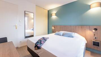 A hotel room with a bed, a mirror, a bedside table, and a wall-mounted lamp.