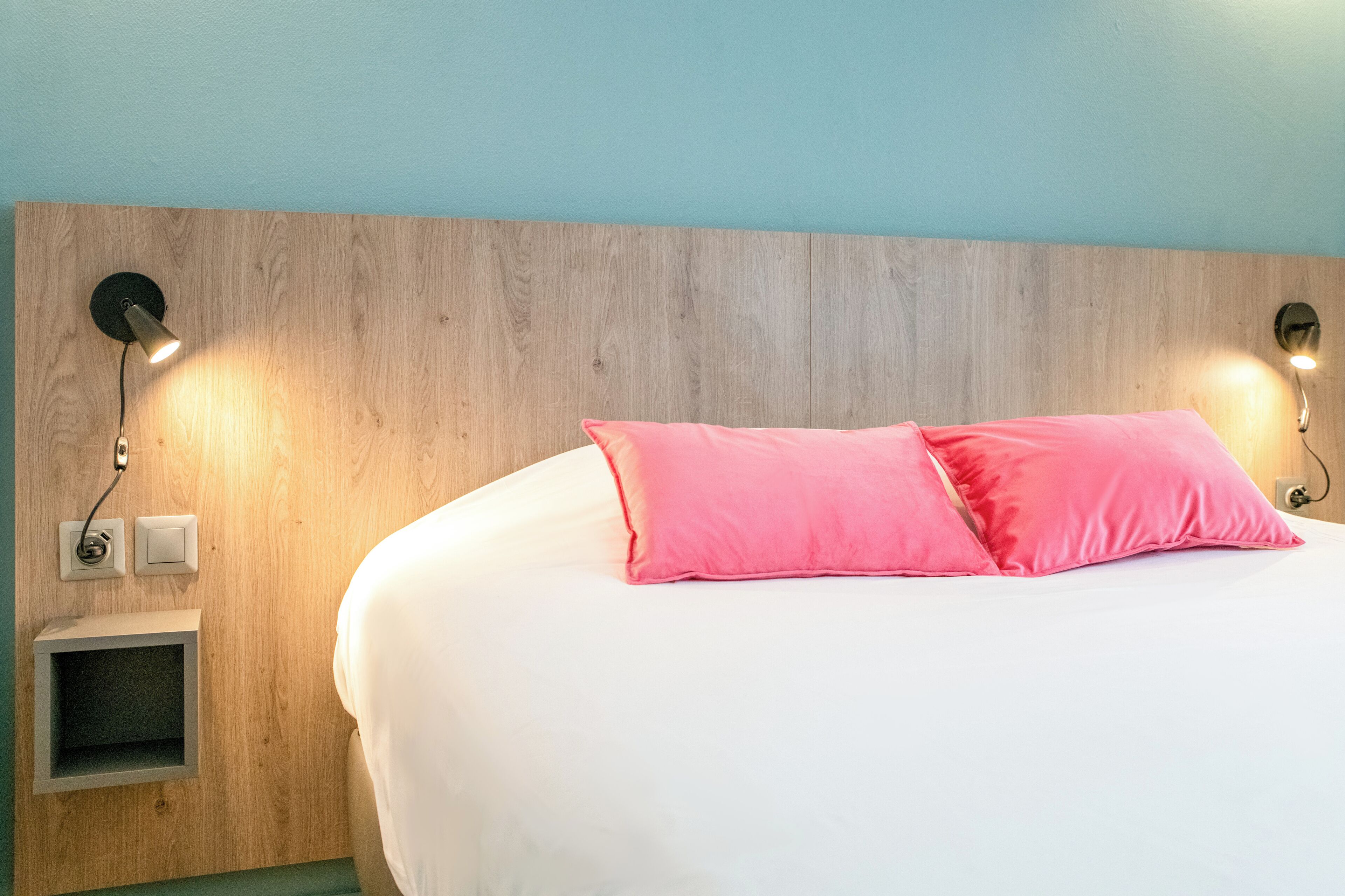 Quarto com cabeceira de madeira, duas luminárias de mesa, cama branca com almofadas cor-de-rosa e um pequeno aparador.