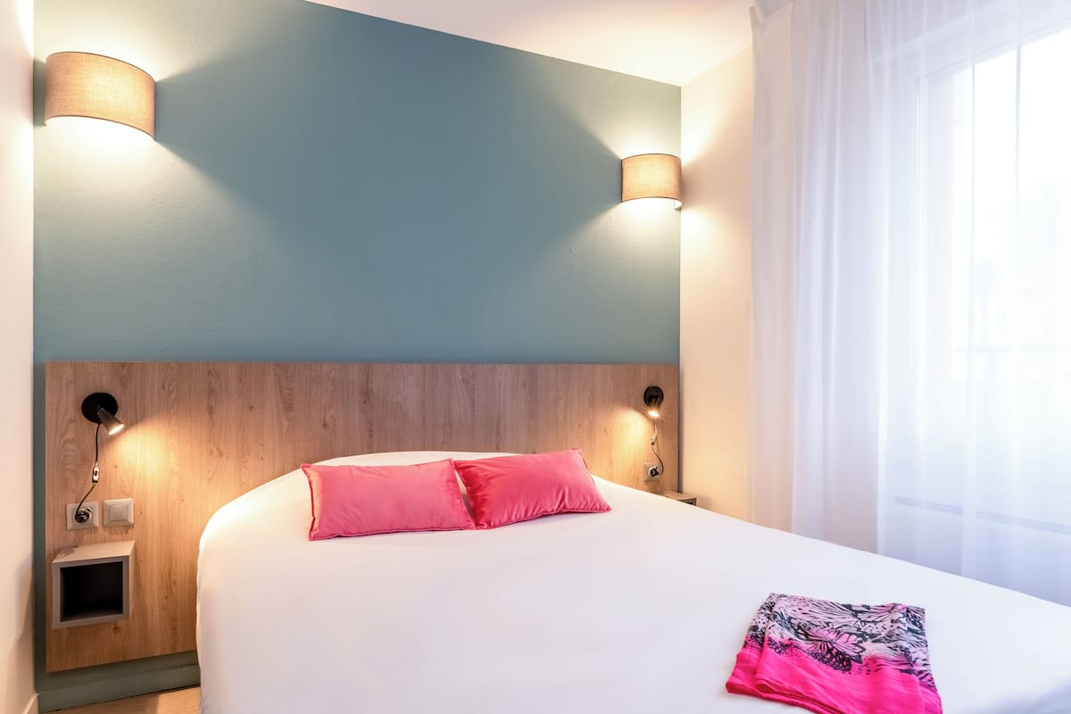 A hotel room with a wooden headboard, two wall-mounted lamps, a bed with white bedding and pink pillows, a small bedside table with a lamp, and a window with sheer curtains.