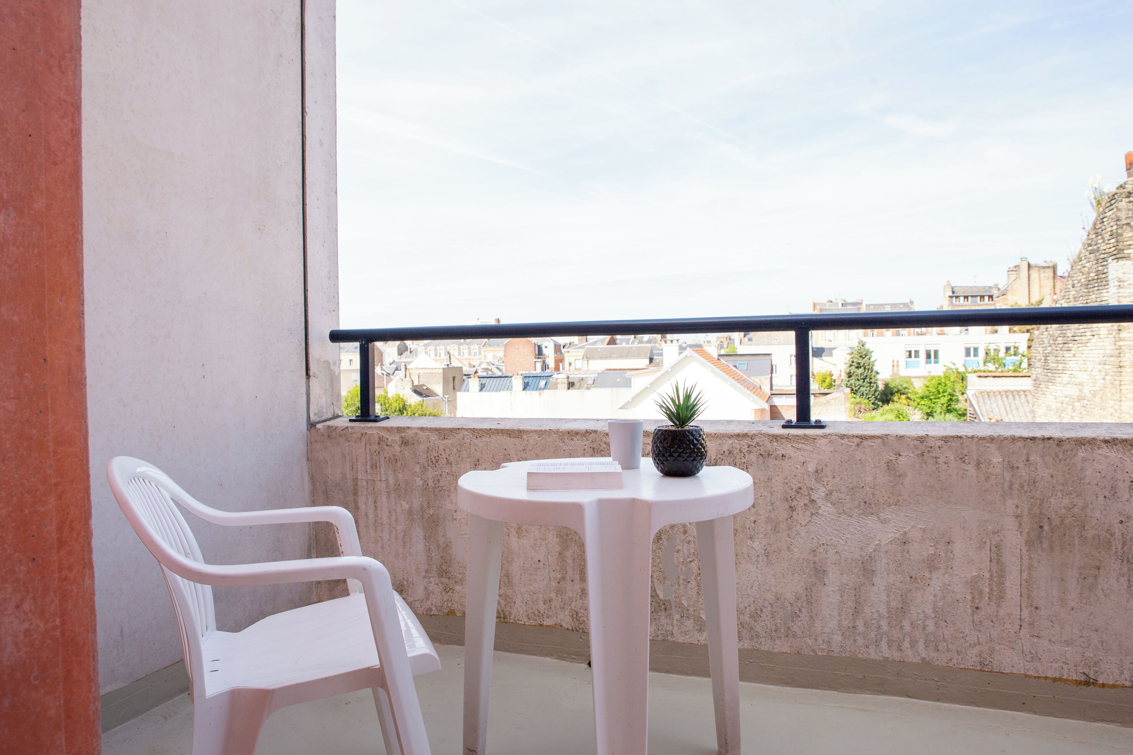 Un balcón con una silla de plástico blanco, una pequeña mesa redonda con una planta en maceta y vistas a edificios al fondo.