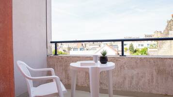 Un balcón con una silla de plástico blanco, una pequeña mesa redonda con una planta en maceta y vistas a edificios al fondo.