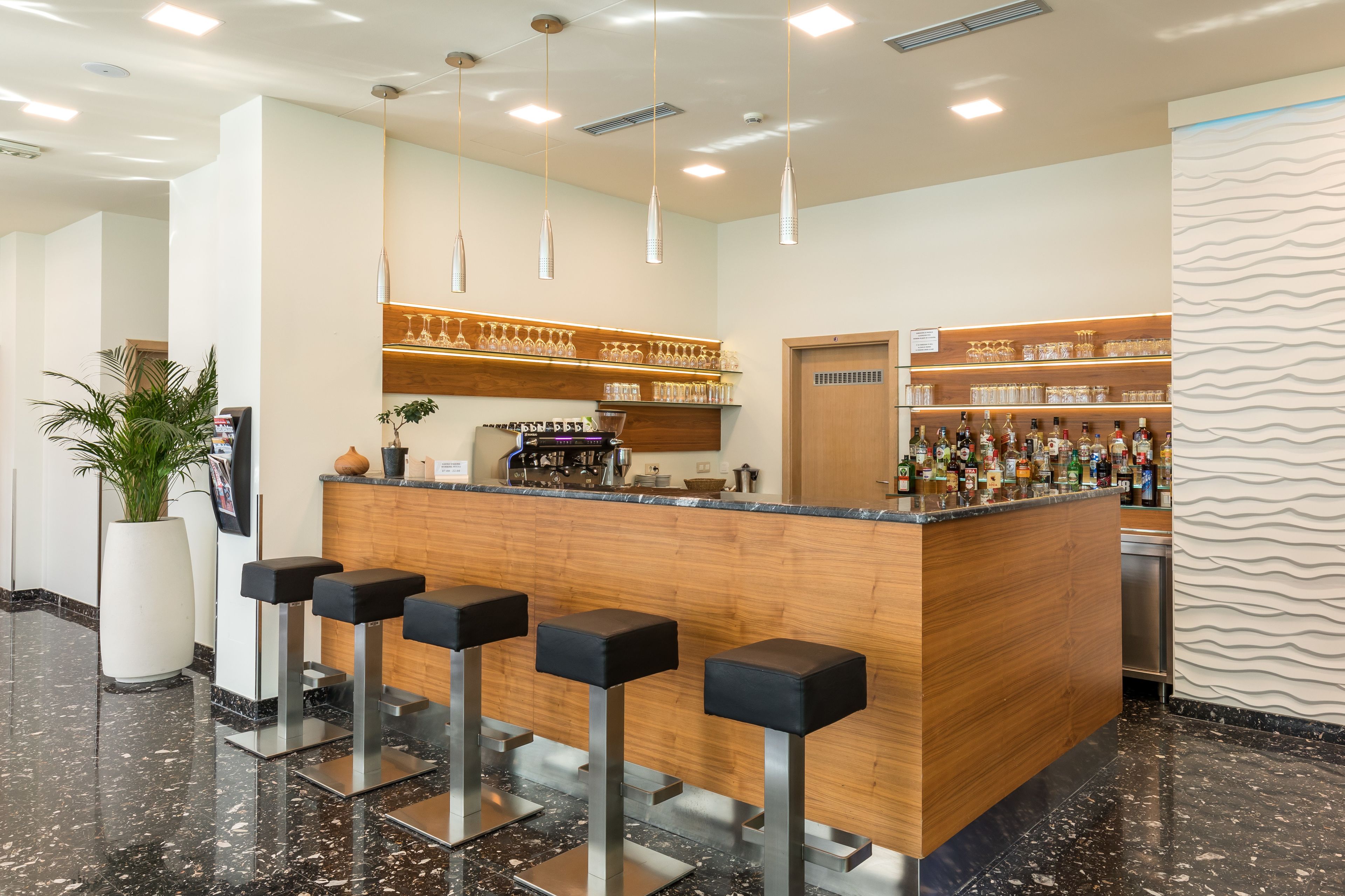 A bar area with a wooden counter, black bar stools, and shelves of bottles and glasses.