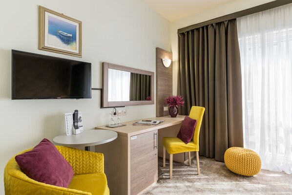 A hotel room with a flat-screen TV, a desk with a mirror, a yellow armchair, a yellow ottoman, and a large window with curtains.