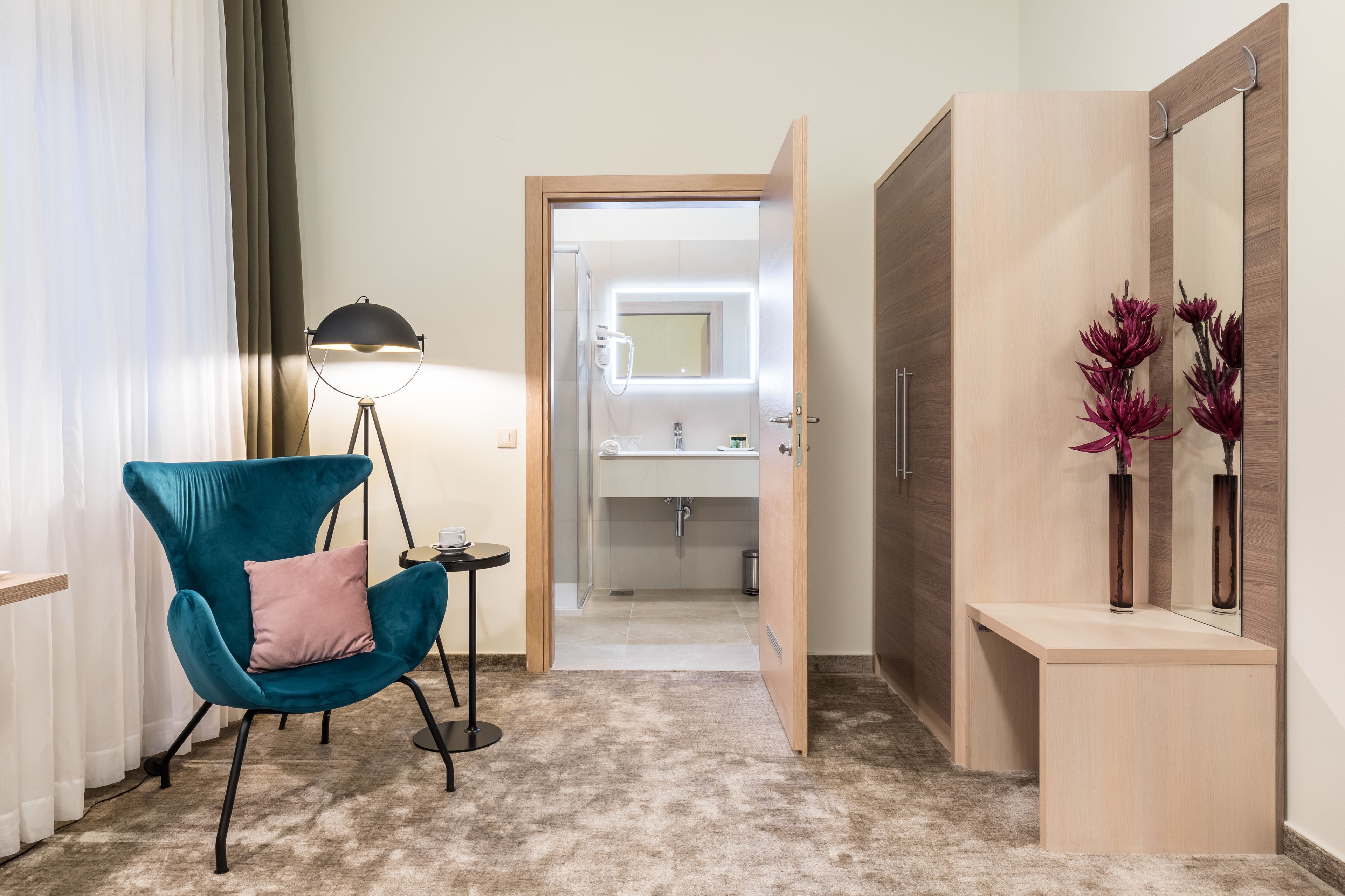 Una habitación de hotel con un sillón verde azulado, una mesita redonda pequeña, una lámpara de pie, un tocador con espejo y un armario de madera con flores decorativas.