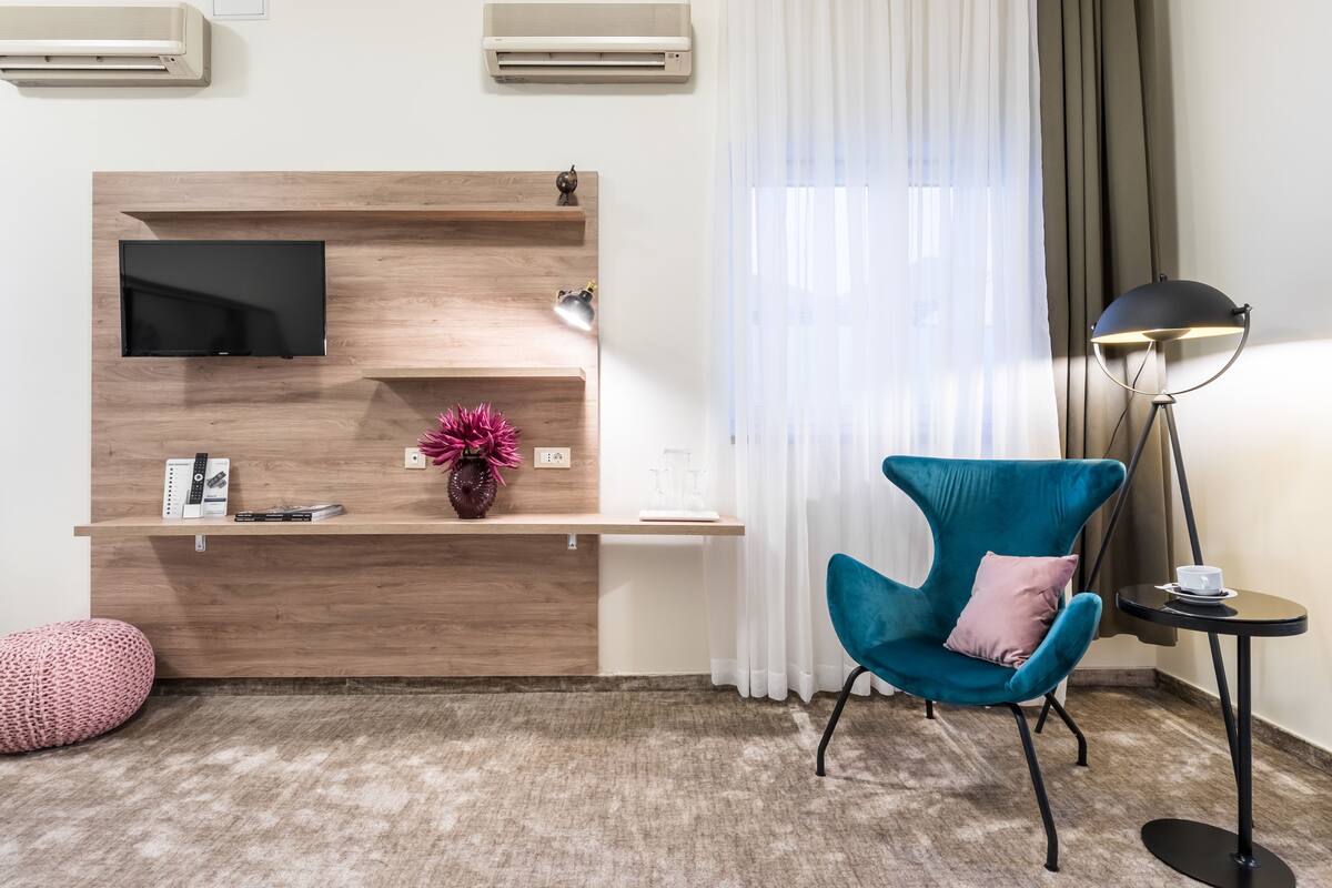 A modern hotel room with a wooden console table, a flat-screen TV, a teal armchair, a small side table with a cup, and a floor lamp.