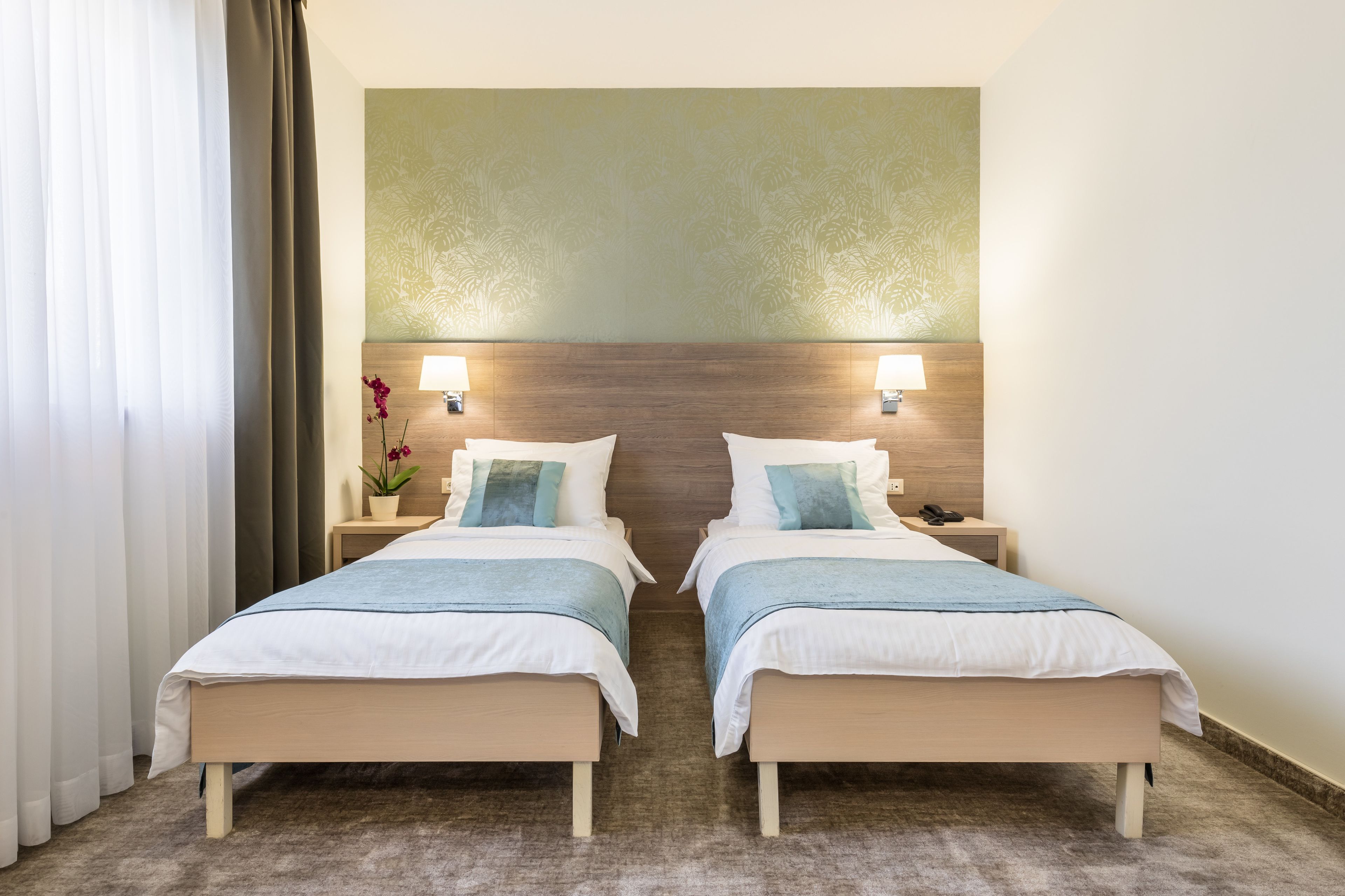 Quarto de hotel com duas camas de solteiro, cabeceira de madeira, mesa de cabeceira com abajur e um vaso de planta.