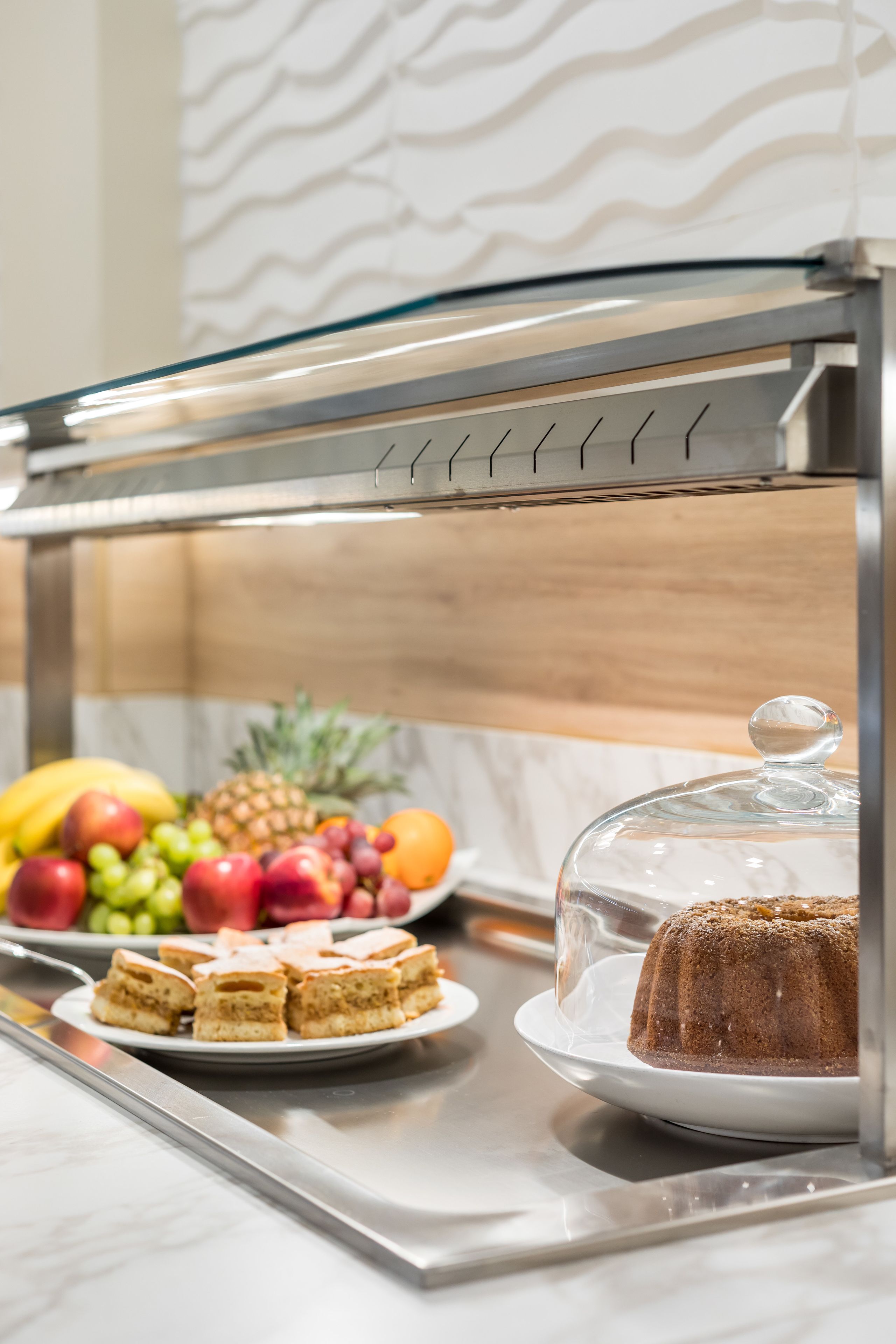 A buffet setup with a glass-covered cake, sliced pastries, and a variety of fruits.
