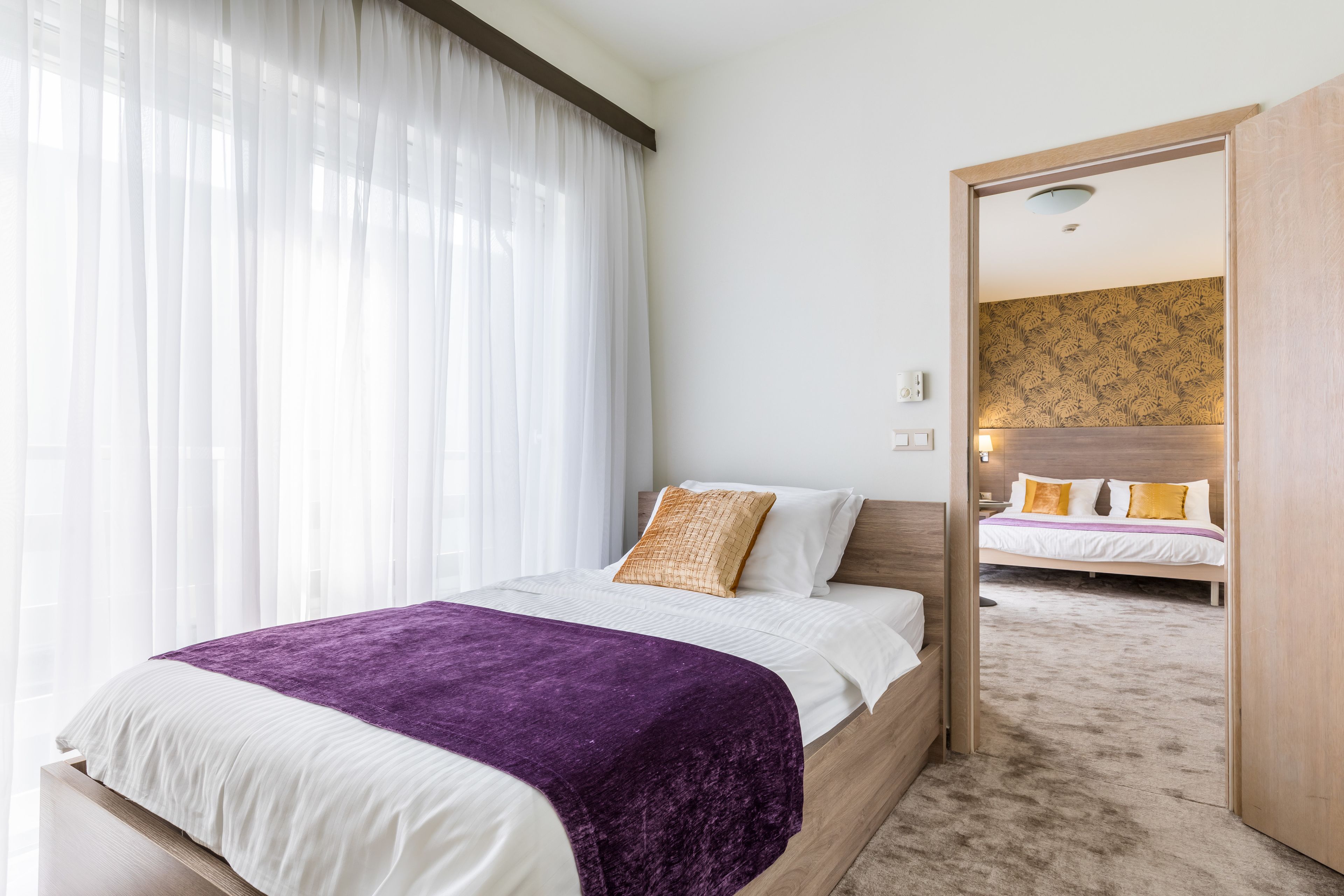A hotel room with a bed, a purple blanket, white bedding, a wooden headboard, and a view through an open door to another room with a patterned wall.