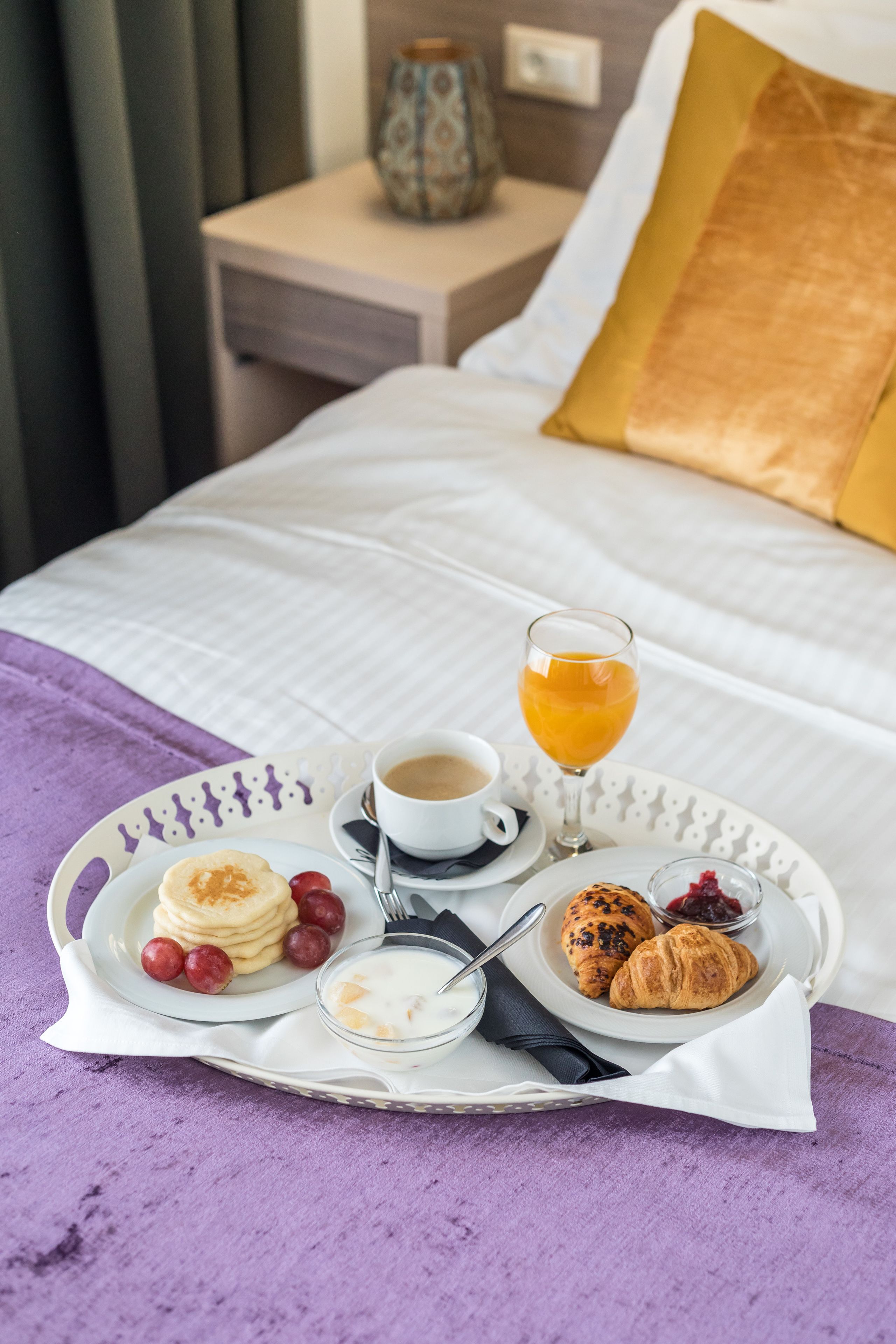 A neatly arranged breakfast tray with pancakes, croissants, and a glass of orange juice on a bed with a white and gold pillow.