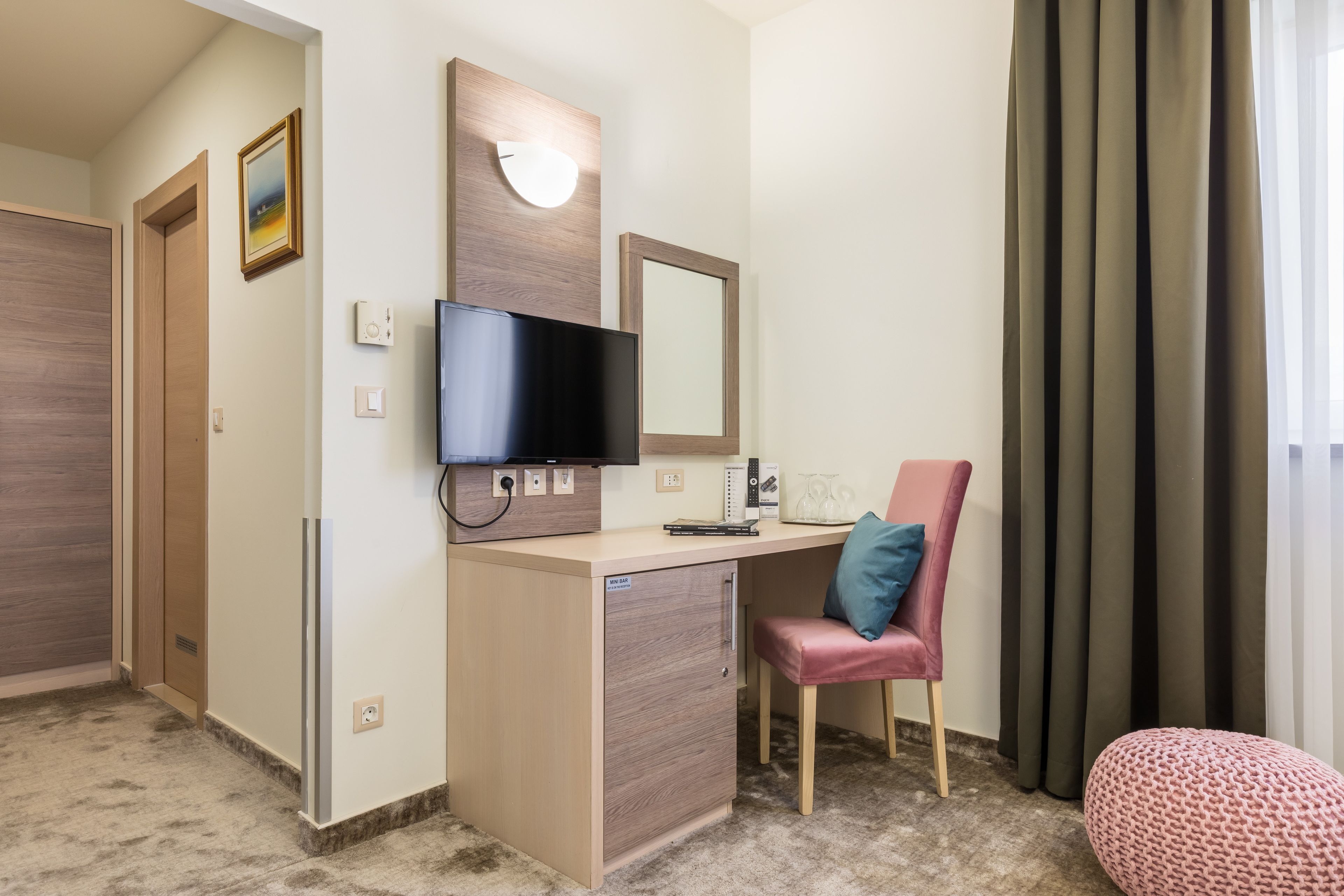 A hotel room with a wooden desk, a television, a mirror, a pink chair, and a window with curtains.