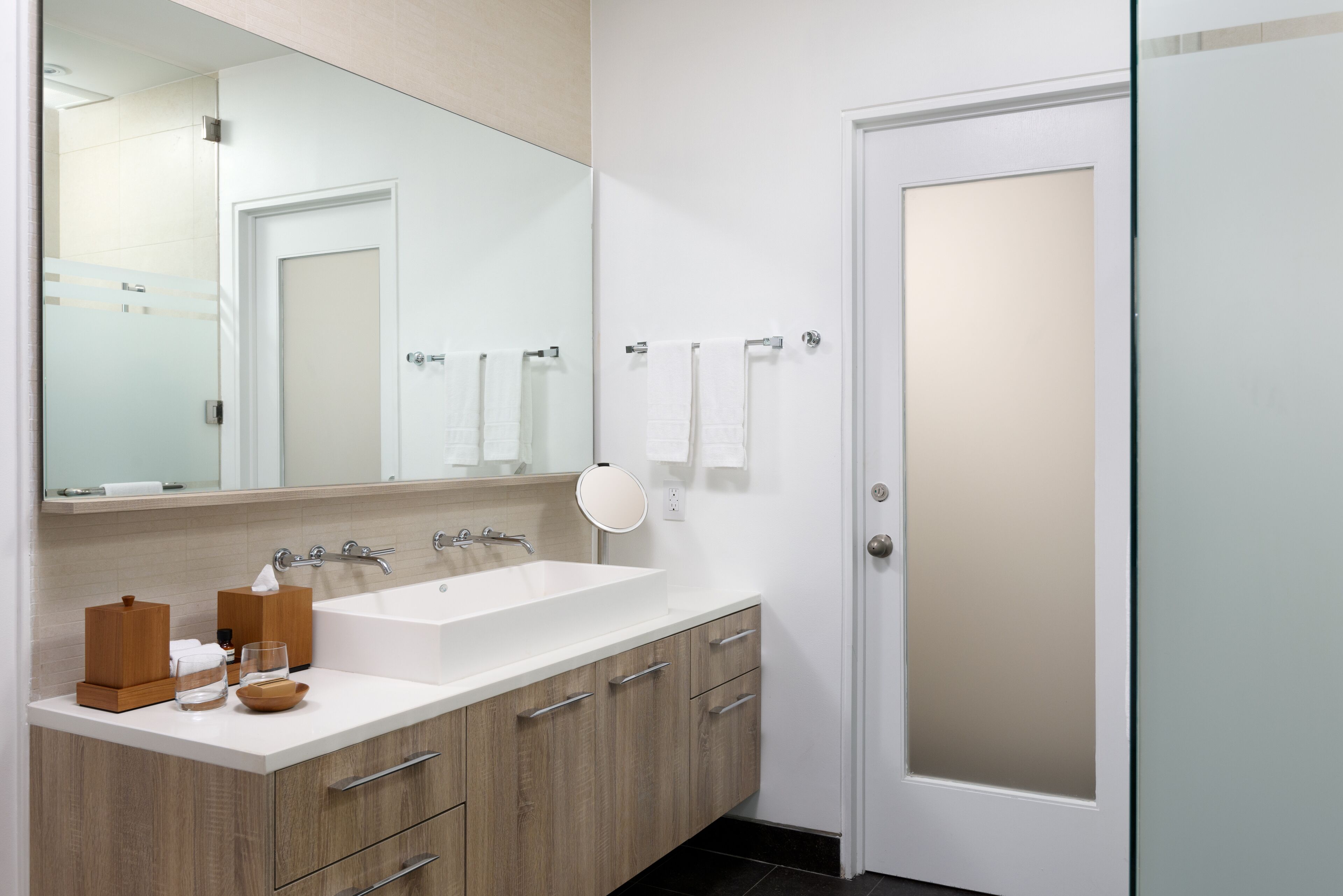 Un baño moderno con un espejo grande, un lavamanos blanco, armarios de madera y una puerta de vidrio esmerilado.