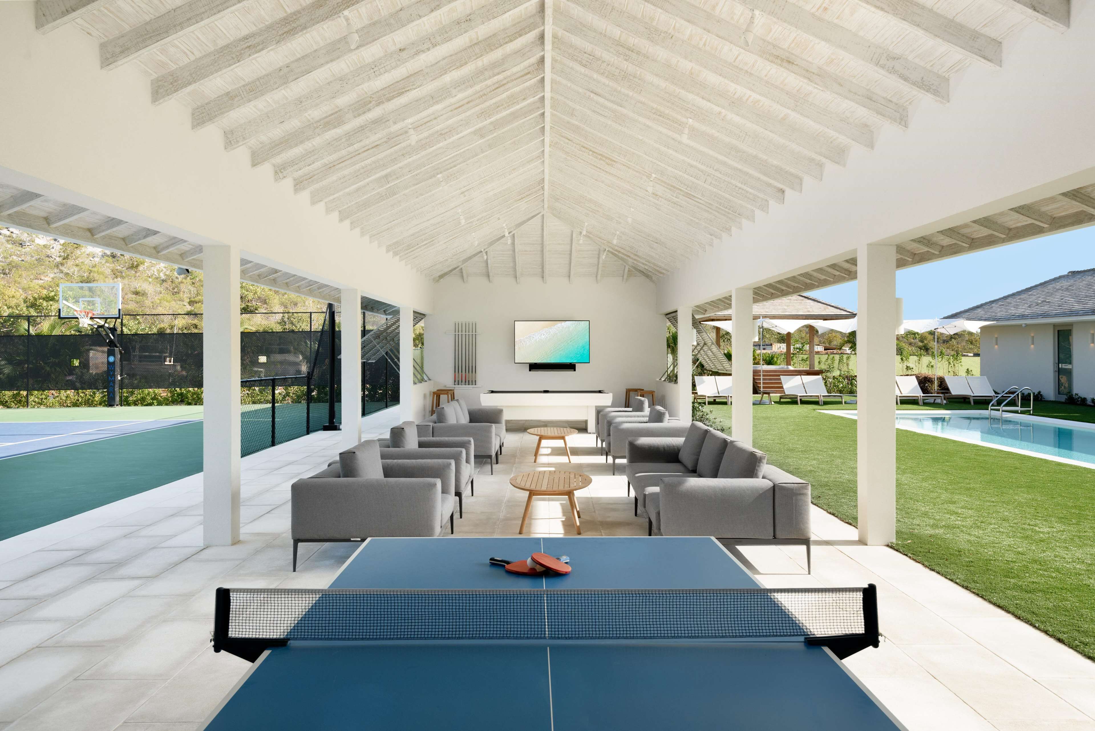 Un área recreativa interior con mesa de ping pong, asientos cómodos, un televisor y una piscina al fondo.