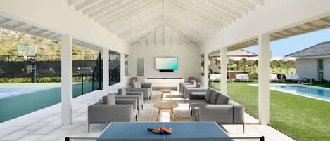 Un área recreativa interior con mesa de ping pong, asientos cómodos, un televisor y una piscina al fondo.