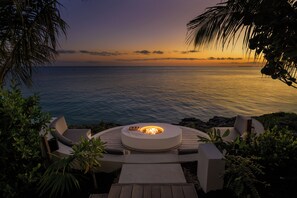 A circular outdoor seating area with a fire pit, surrounded by greenery and overlooking a body of water at sunset.