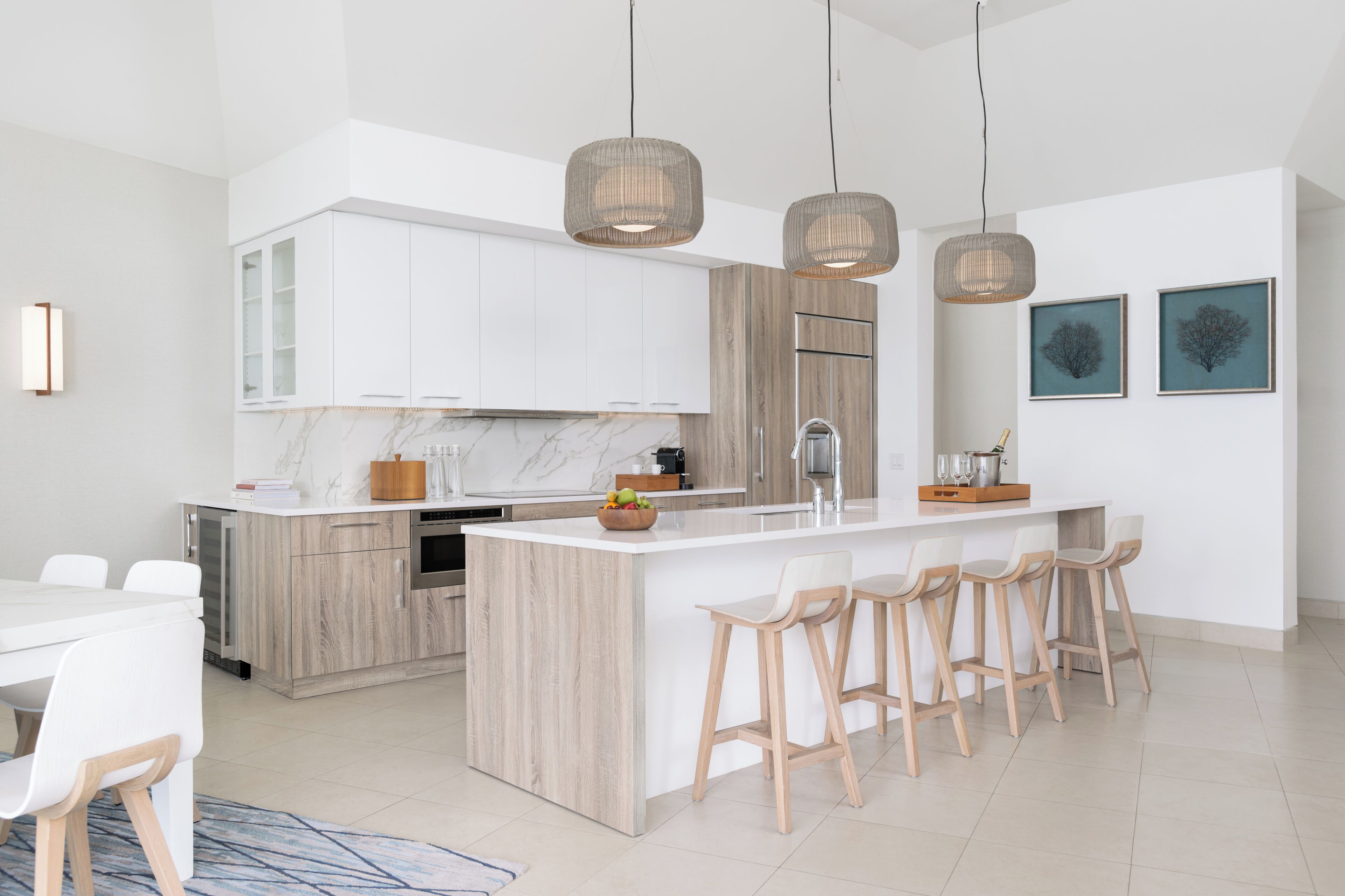 Una cocina moderna con armarios blancos, encimera de mármol y detalles de madera. Hay tres lámparas colgantes, un horno empotrado y una isla grande con taburetes de bar.