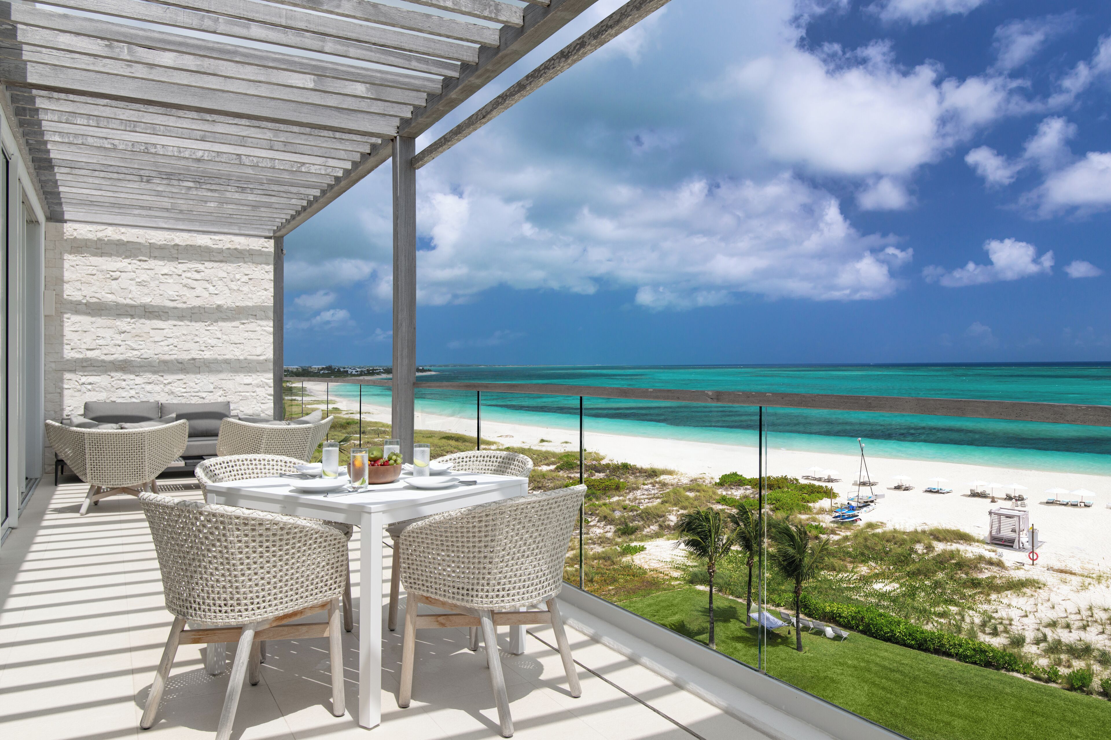 A balcony with a table and chairs overlooking a beach with clear turquoise waters and a white sandy shore.