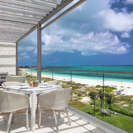 A balcony with a table and chairs overlooking a beach with clear turquoise waters and a white sandy shore.