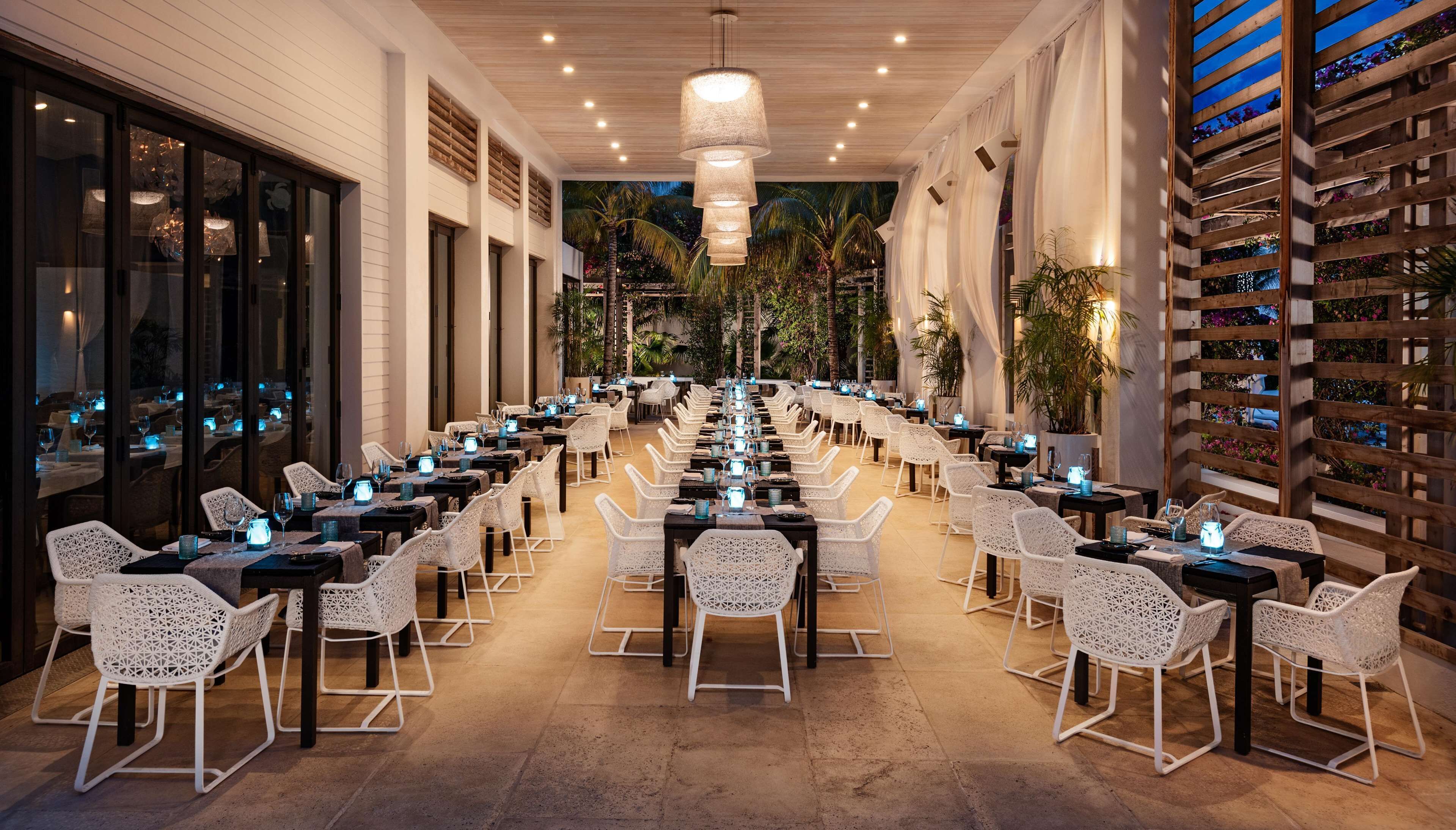 A spacious outdoor dining area with white chairs and tables, a central hanging light fixture, and a backdrop of plants and a wooden fence.