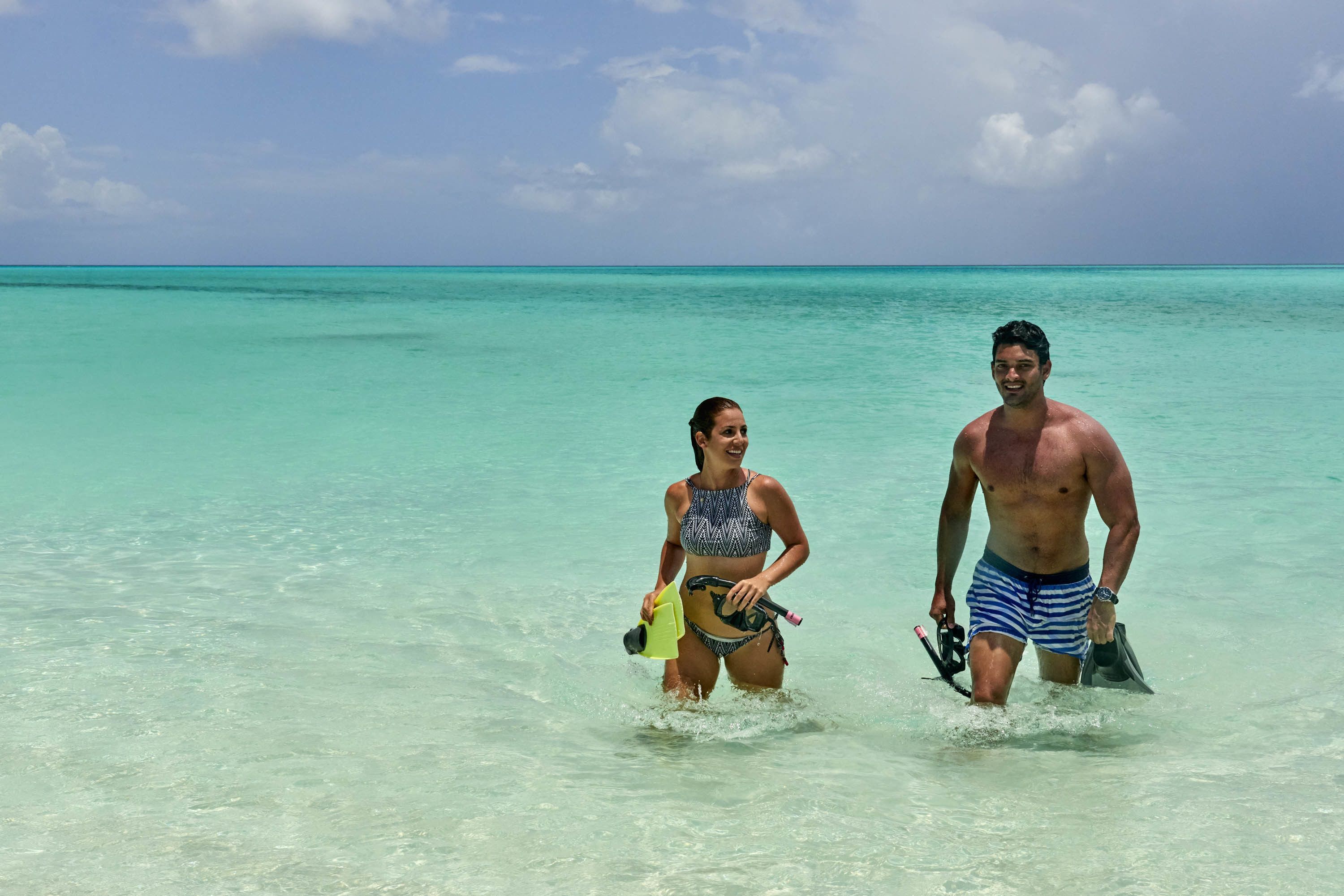 Two people in shallow water, one holding a snorkel and fins, and the other holding a camera.