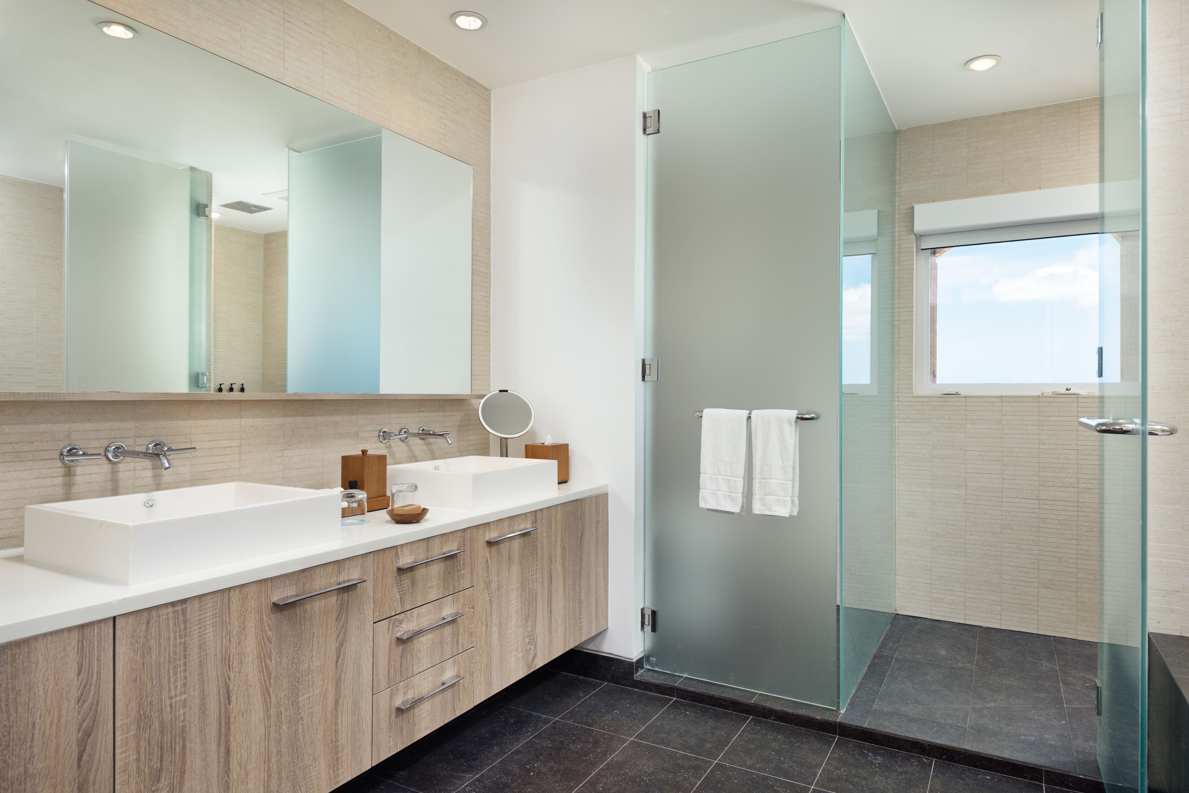 A modern bathroom with a large mirror, two sinks, wooden cabinets, and a glass-enclosed shower.