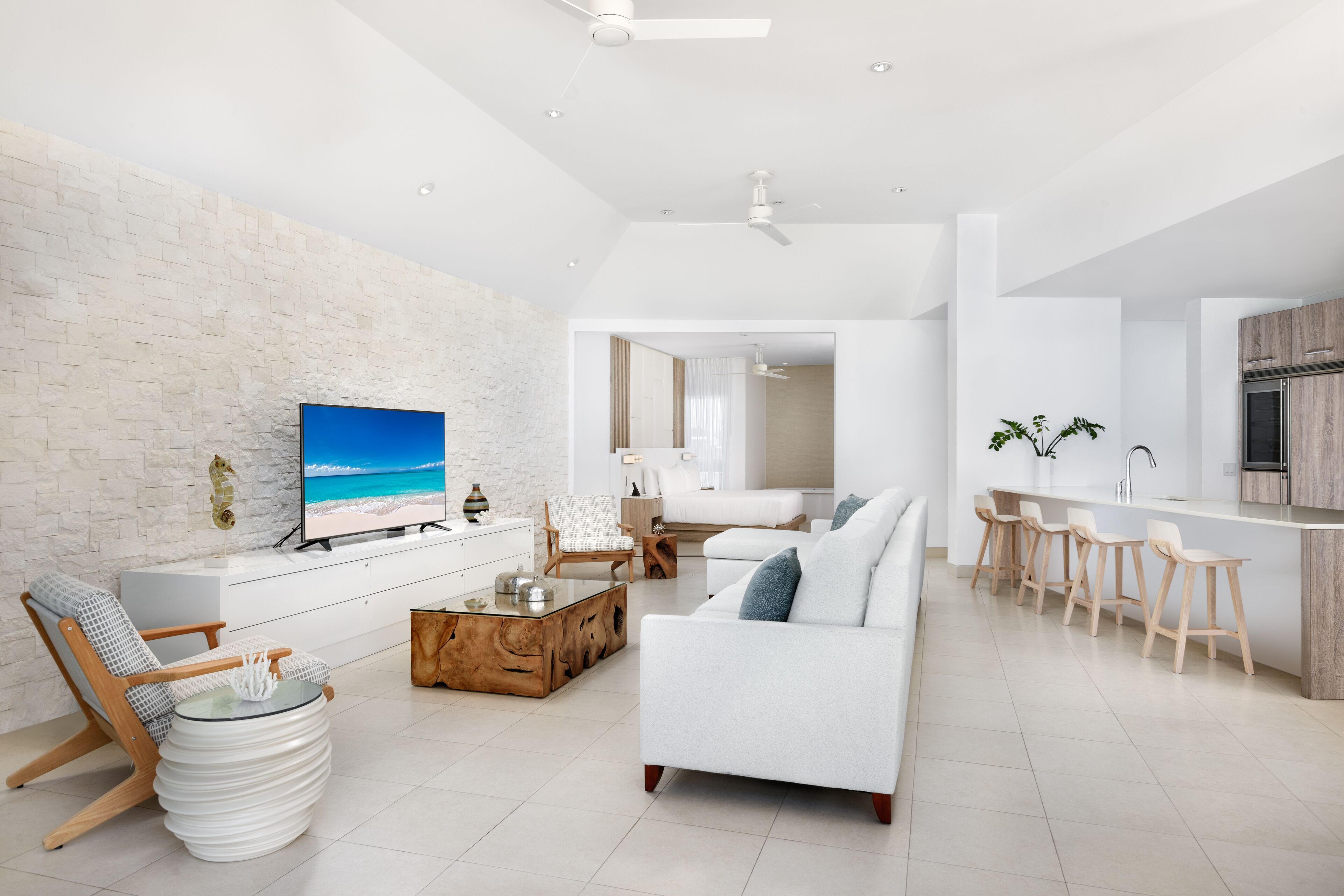 Un salón moderno con un sofá blanco, una mesa de centro y un televisor que muestra una escena de playa. Hay una zona de cocina con taburetes de bar y una encimera blanca.