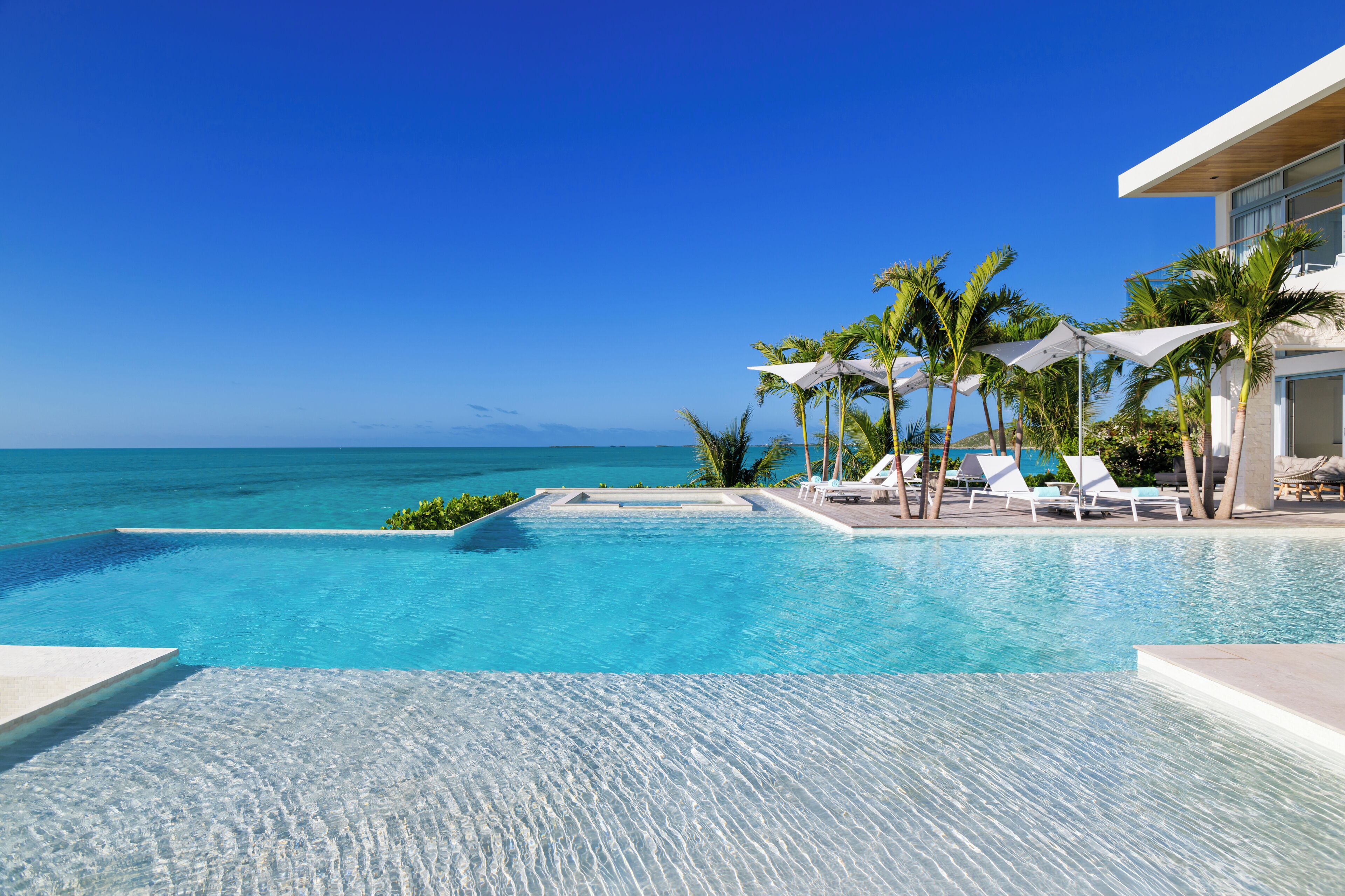 A clear blue pool with white lounge chairs and umbrellas, overlooking the ocean.