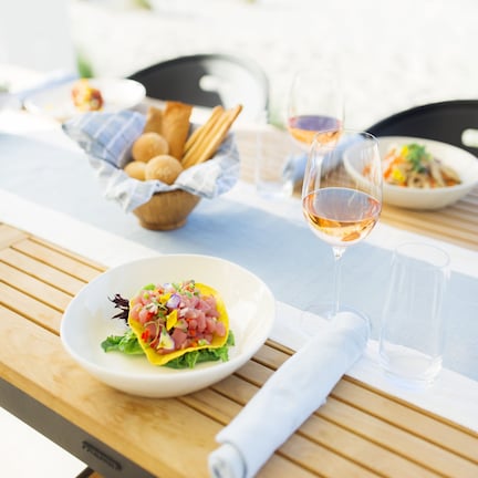 A dining table with a plate of food, a glass of wine, and a bowl of bread on a wooden table outdoors.