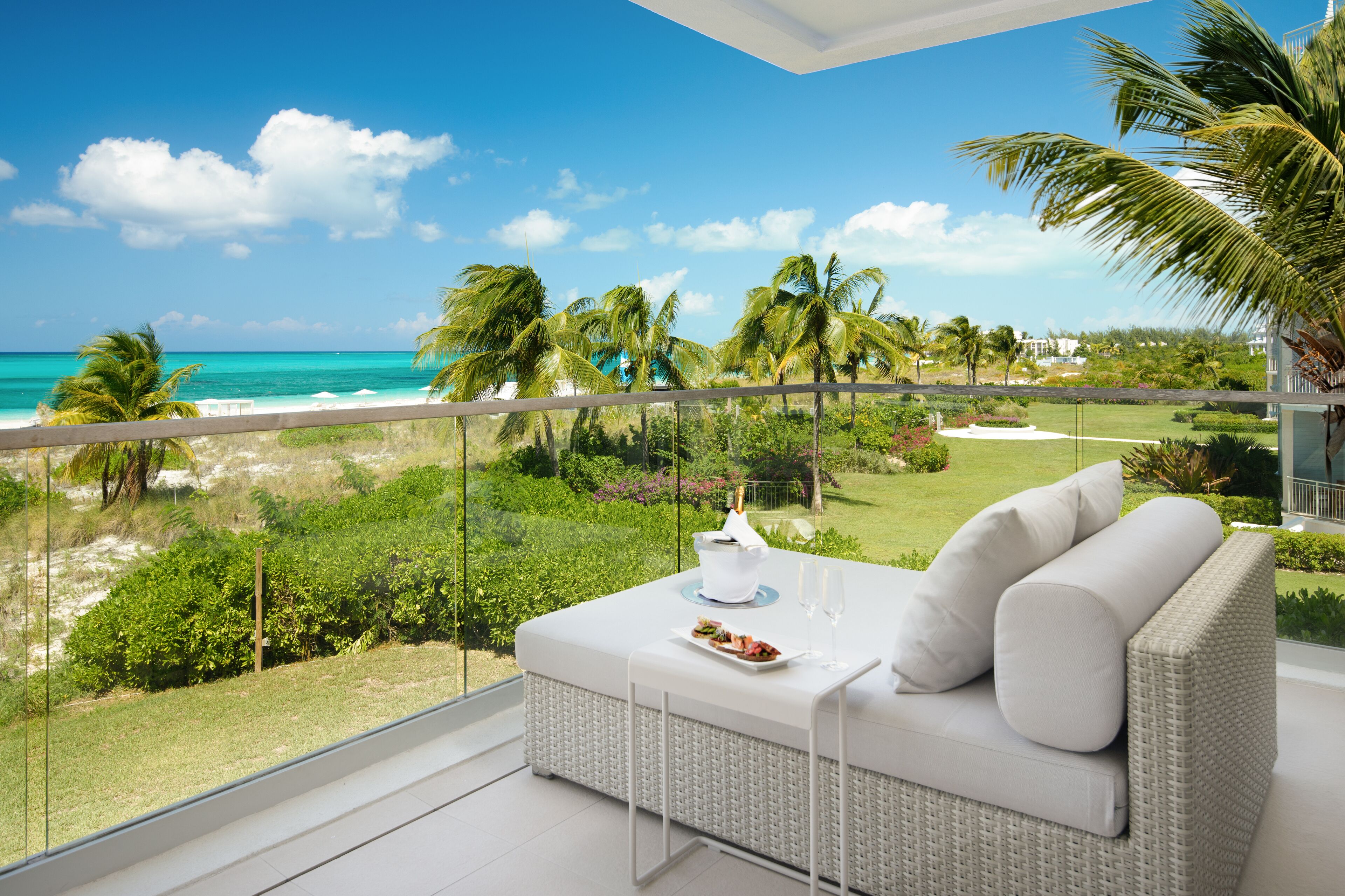 A balcony with a white wicker sofa, a small table with a plate of food and two glasses of water, and a view of the ocean and palm trees.