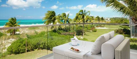 A balcony with a white wicker sofa, a small table with a plate of food and two glasses of water, and a view of the ocean and palm trees.