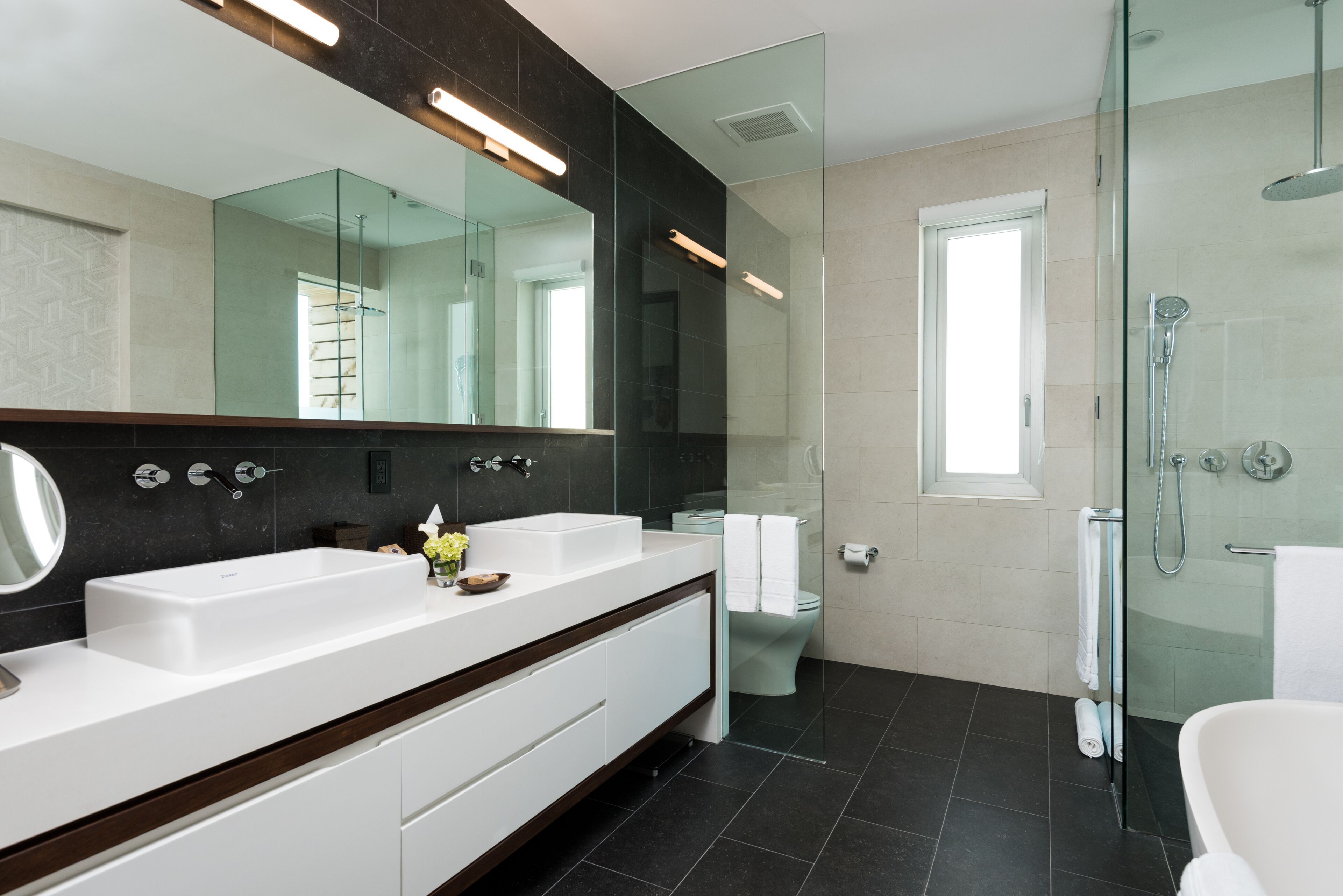 A modern bathroom with a large mirror, two sinks, a glass shower enclosure, and a toilet.