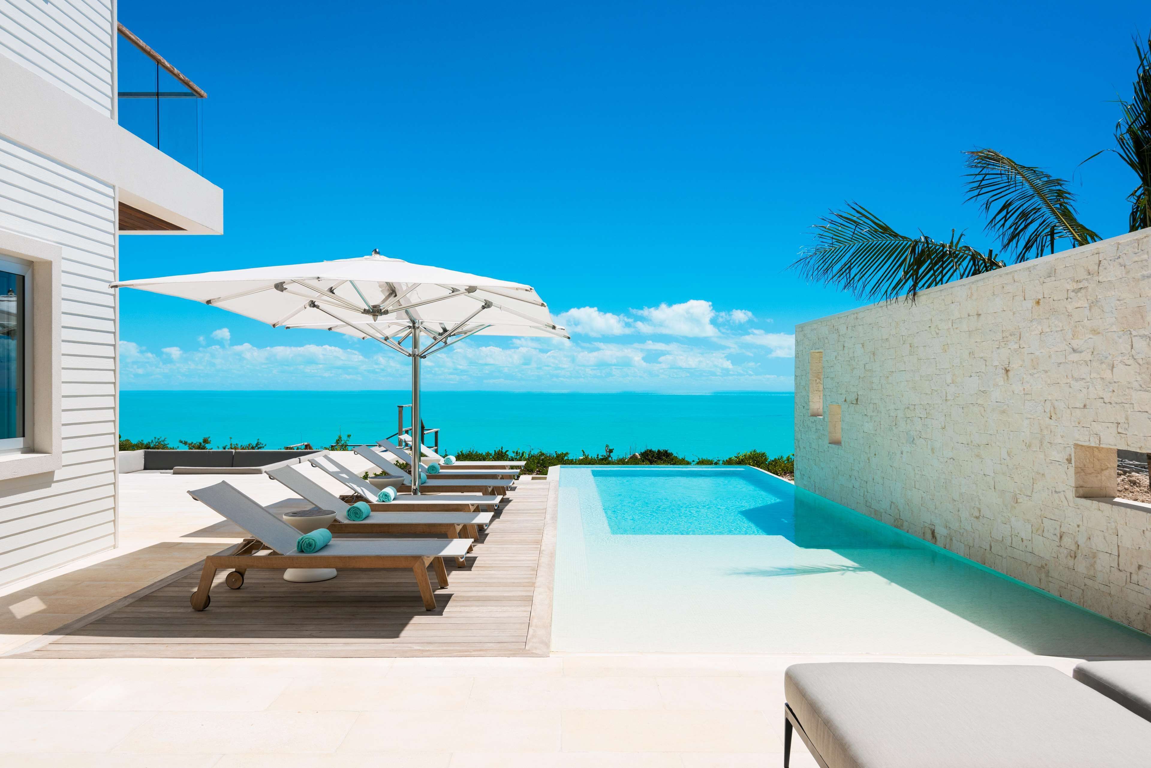 Un área de piscina moderna al aire libre con sillones de descanso y sombrillas, con vista a un mar azul y transparente.