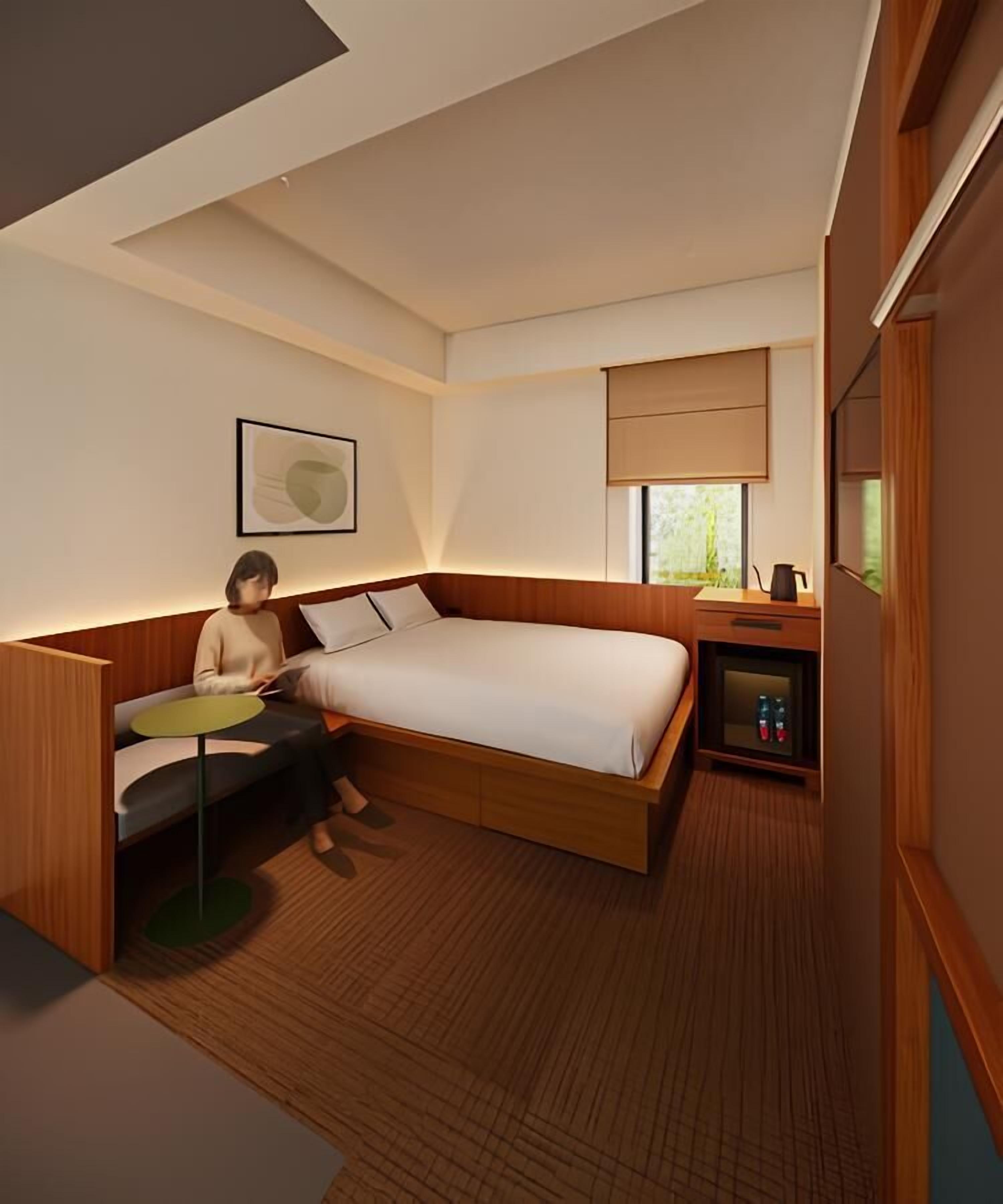 A modern hotel room with a bed, a desk, a small table, a nightstand, and a window with blinds.