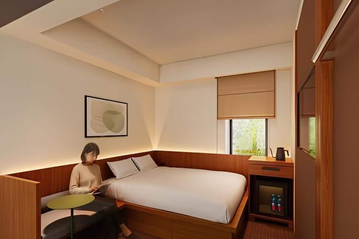 Une chambre d’hôtel moderne équipée d’un lit, d’un bureau, d’une petite table, d’une table de chevet et d’une fenêtre avec des stores.