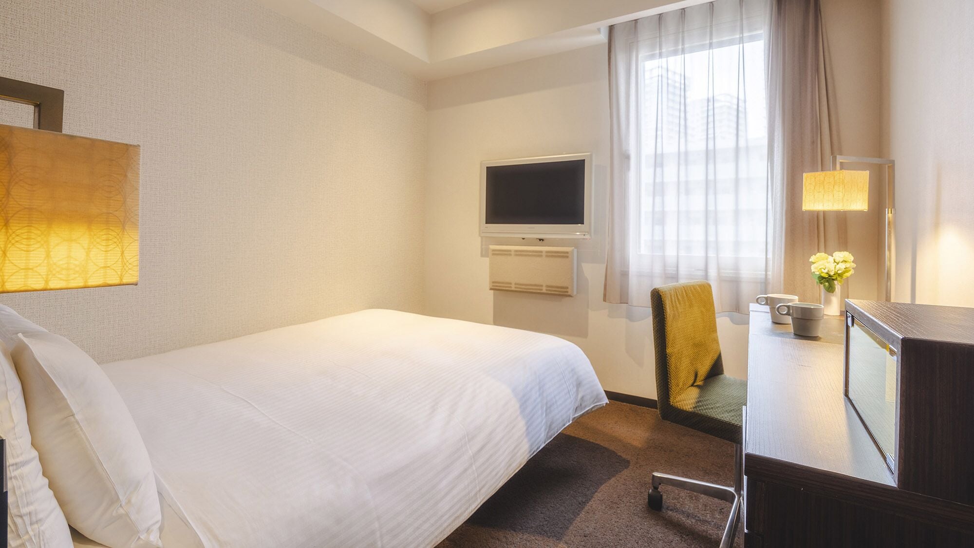 Habitación de hotel con cama, escritorio con lámpara, una silla, televisión y ventana con cortinas.