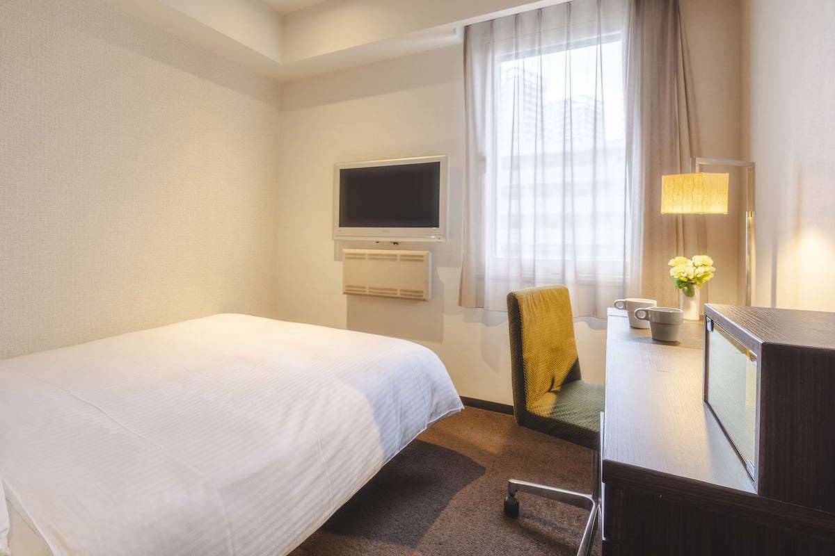 Habitación de hotel con cama, escritorio con lámpara, una silla, televisión y ventana con cortinas.