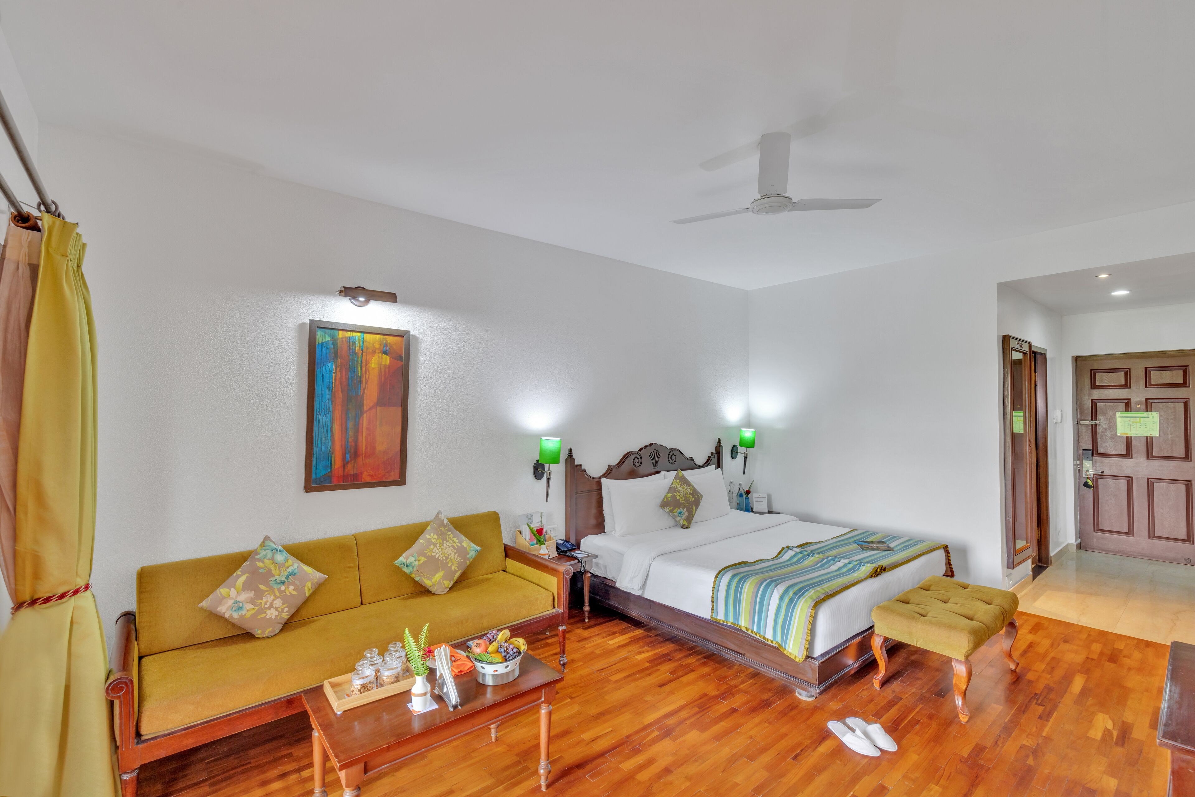 A hotel room with a bed, a sofa, a small table, and a ceiling fan.