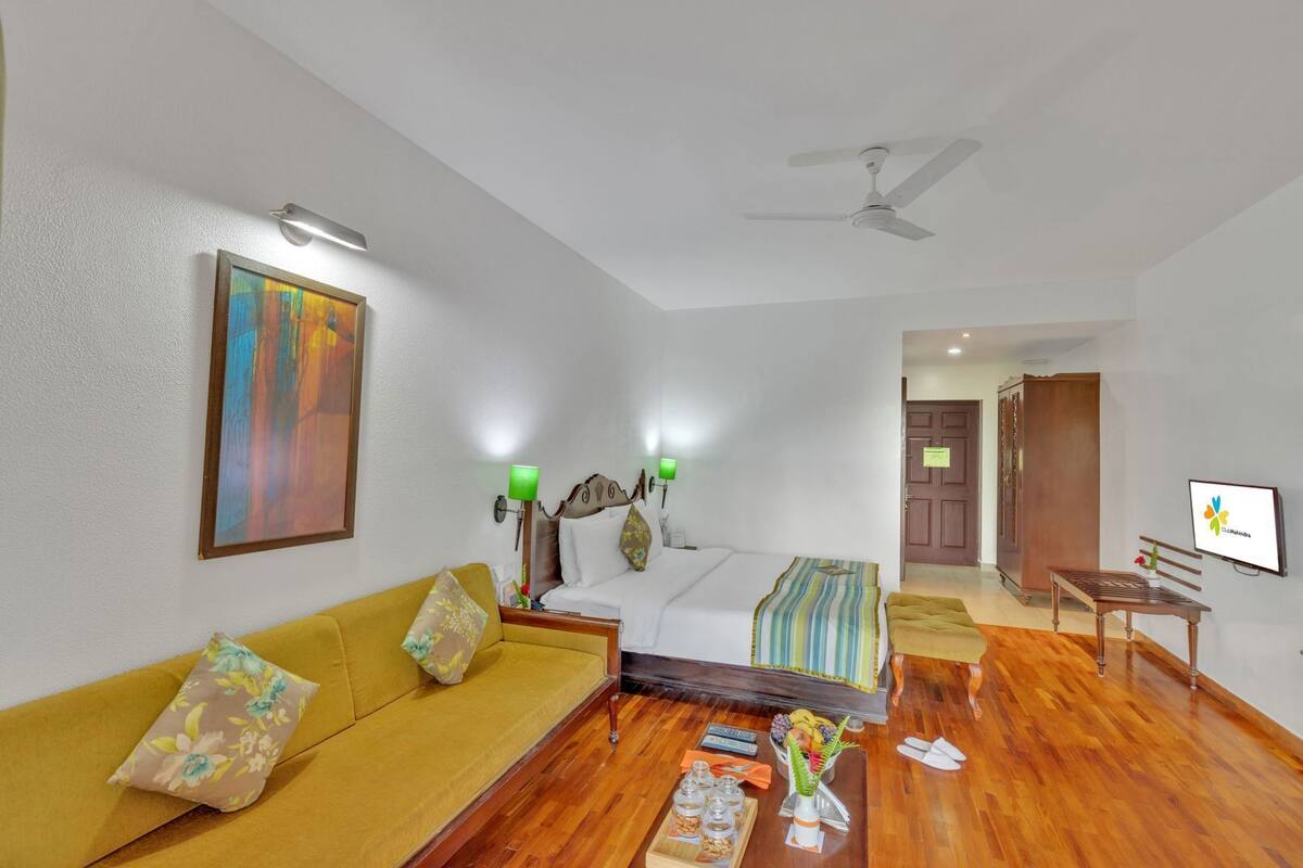 A hotel room with a bed, a sofa, a TV, a ceiling fan, and a painting on the wall.