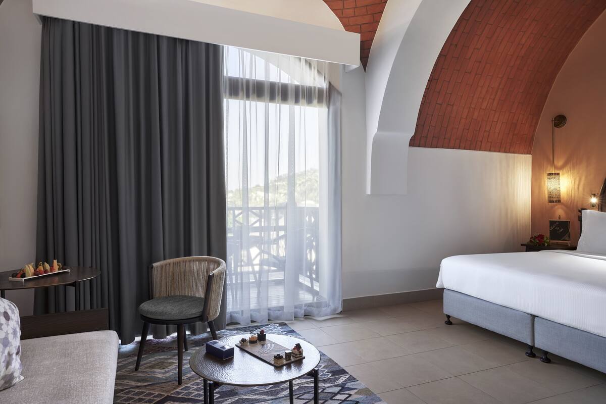 A hotel room with a bed, a chair, a small table with a tray of fruit, and a view of the outdoors.