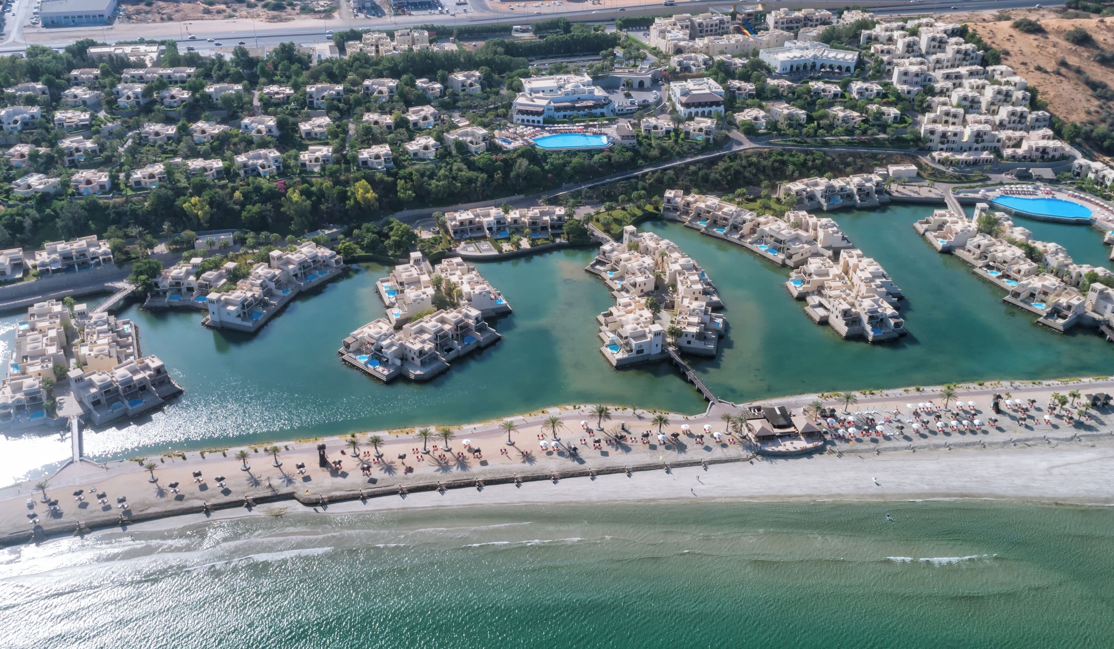 An aerial view of a coastal resort with multiple villas connected by walkways over water, a sandy beach, and a marina.