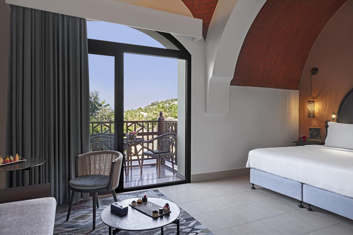 A hotel room with a bed, a chair, a small table with items, a balcony with chairs and a table, and a view of greenery.