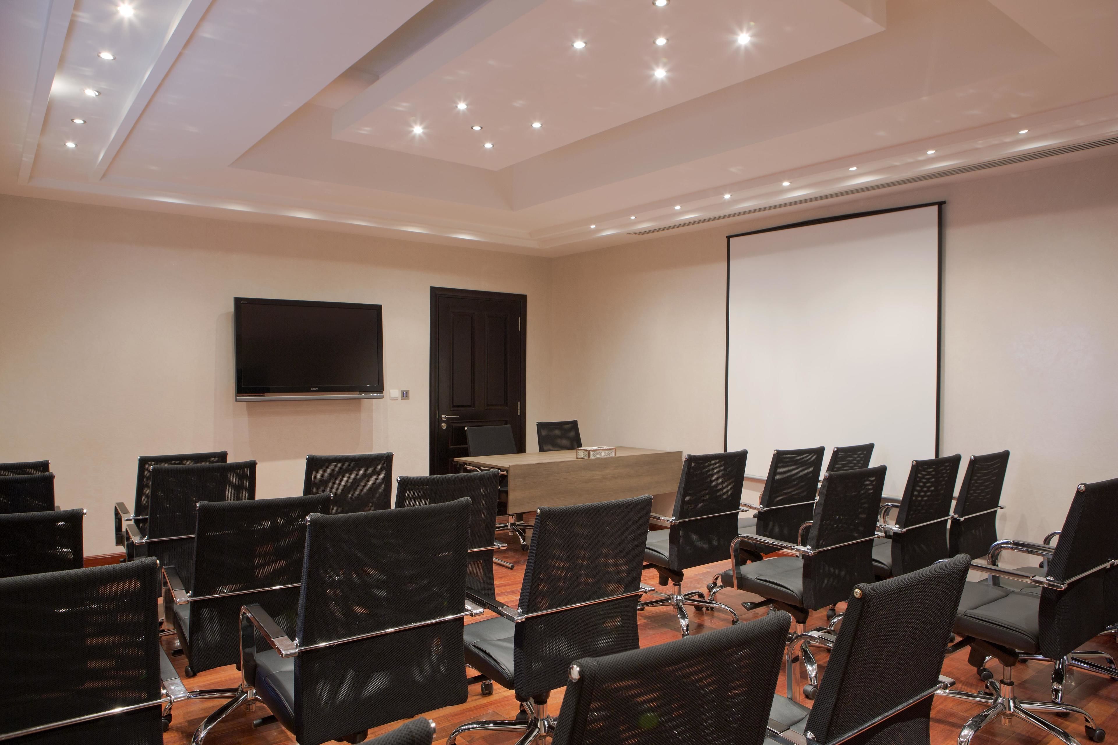 A conference room with a large table, black chairs, a flat-screen TV, and a white projection screen.