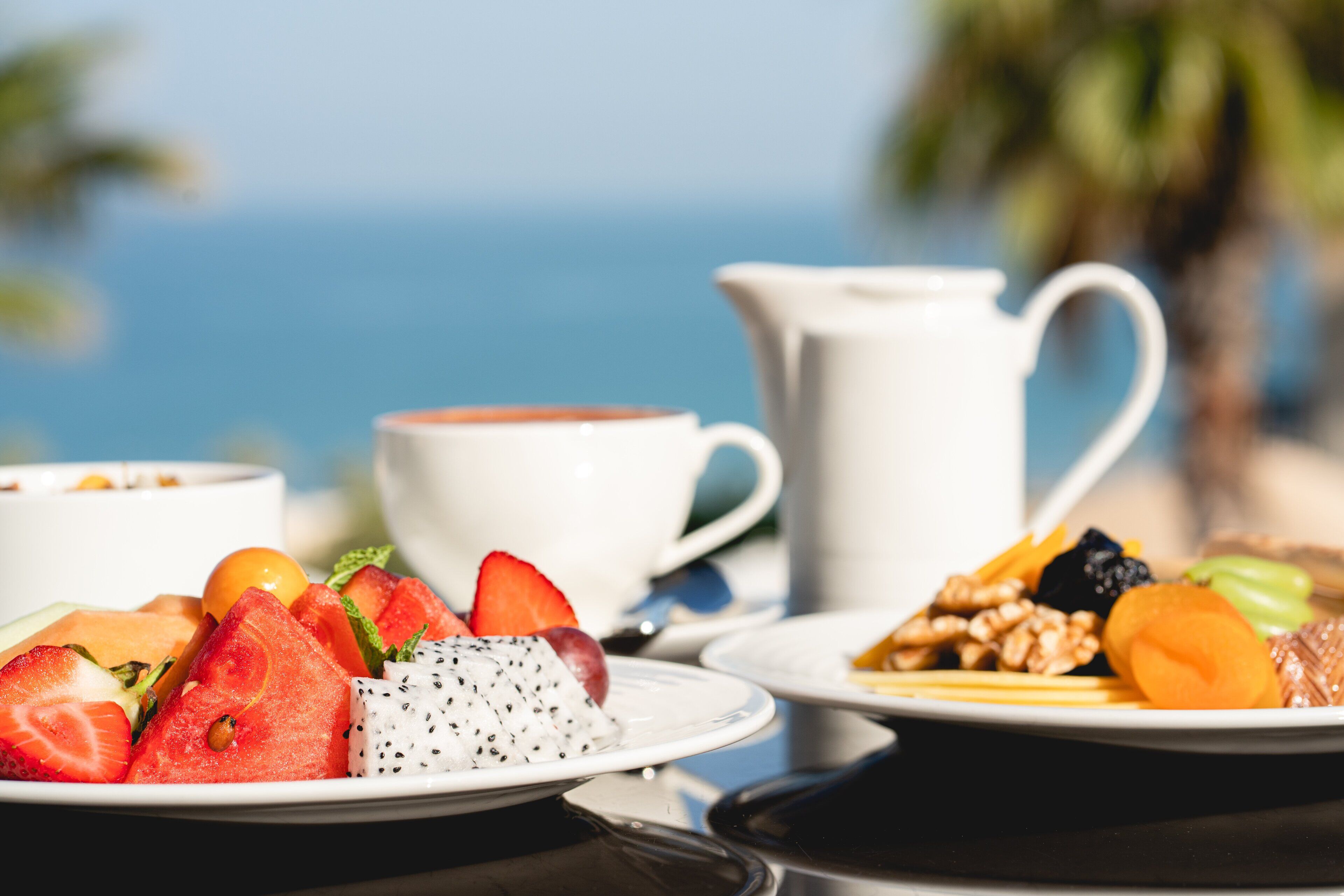Ein Tisch mit Fruchtschalen und einer Kaffeetasse, vor verschwommenem Hintergrund von Strand und Palmen.