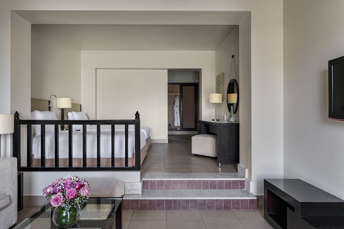 A hotel room with a bed, a sitting area with a table and a vase of flowers, a bathroom with a mirror and sink, and a TV mounted on the wall.