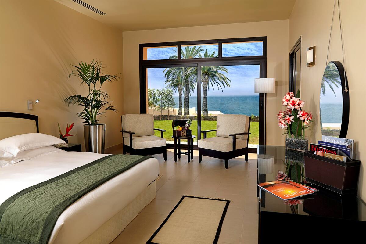 A hotel room with a bed, a desk, a chair, a potted plant, a view of the ocean, and a window with a view of palm trees.