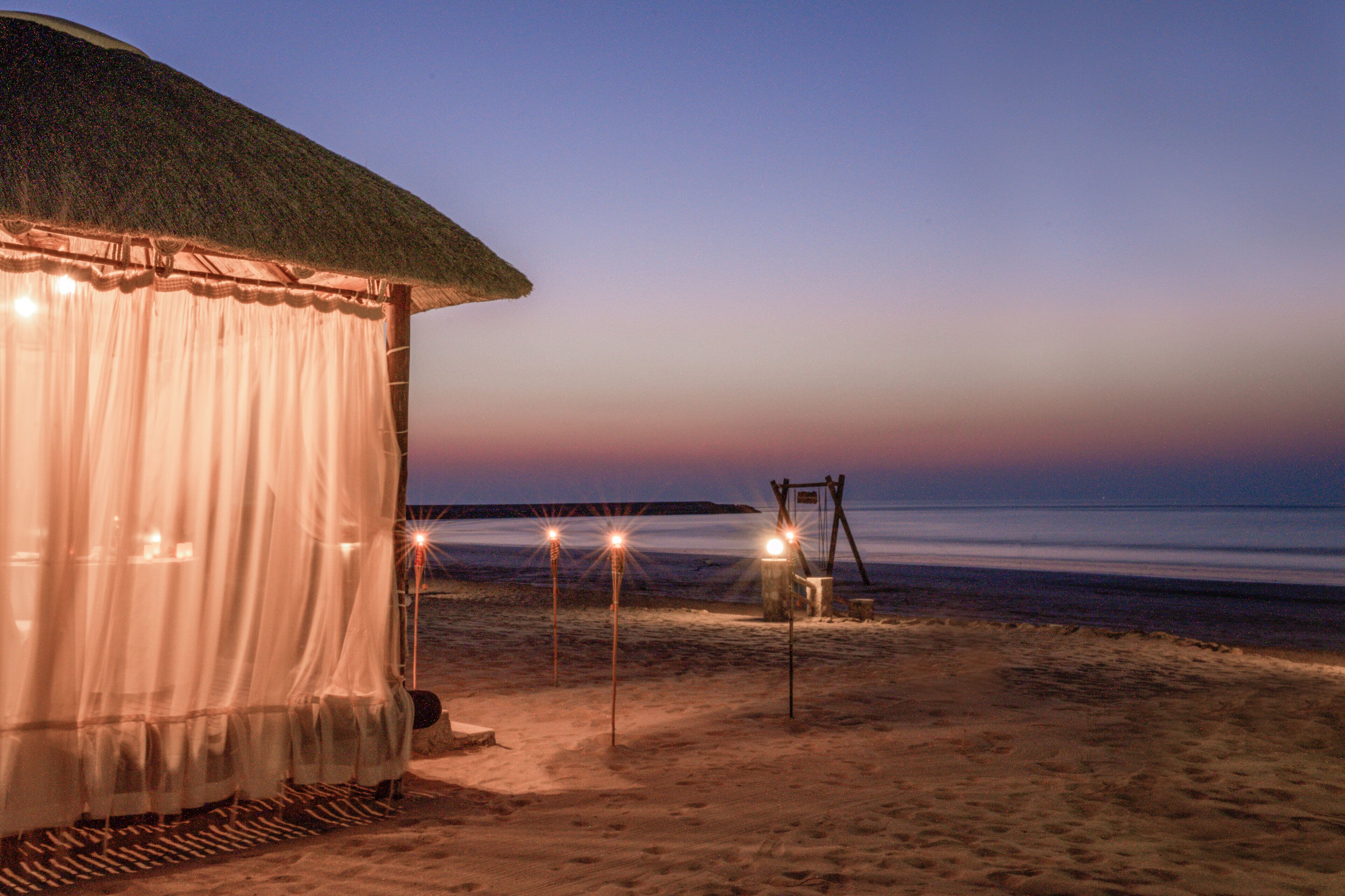 Una cabaña de playa con techo de paja y una cortina blanca. Hay tres luces sobre postes en la arena.