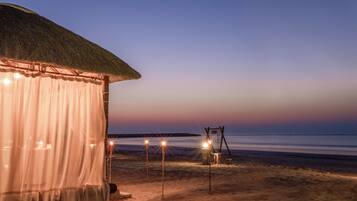 Una cabaña de playa con techo de paja y una cortina blanca. Hay tres luces sobre postes en la arena.